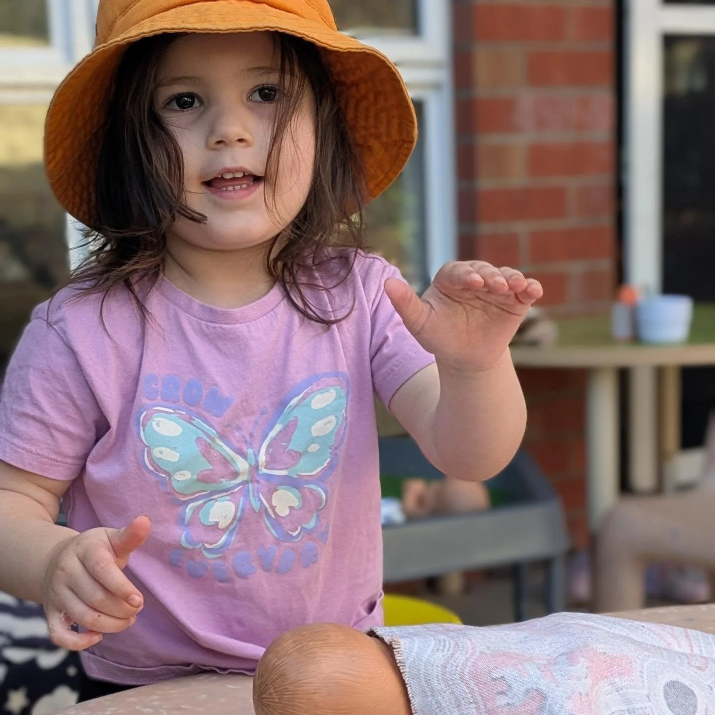 Young girl wearing a pink shirt with a butterfly graphic, orange hat, and brown hair, gesturing with her hand outdoors near a brick wall and table.