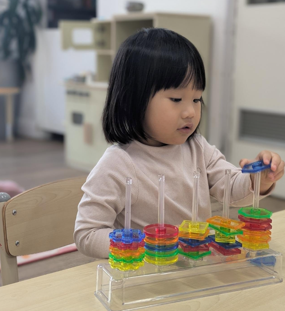 A young girl with black hair playing with colorful stacking rings at a classroom table.