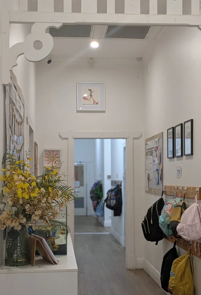 Interior of a hallway or entrance area with a floral arrangement, pictures on the wall, and backpacks hanging on hooks.