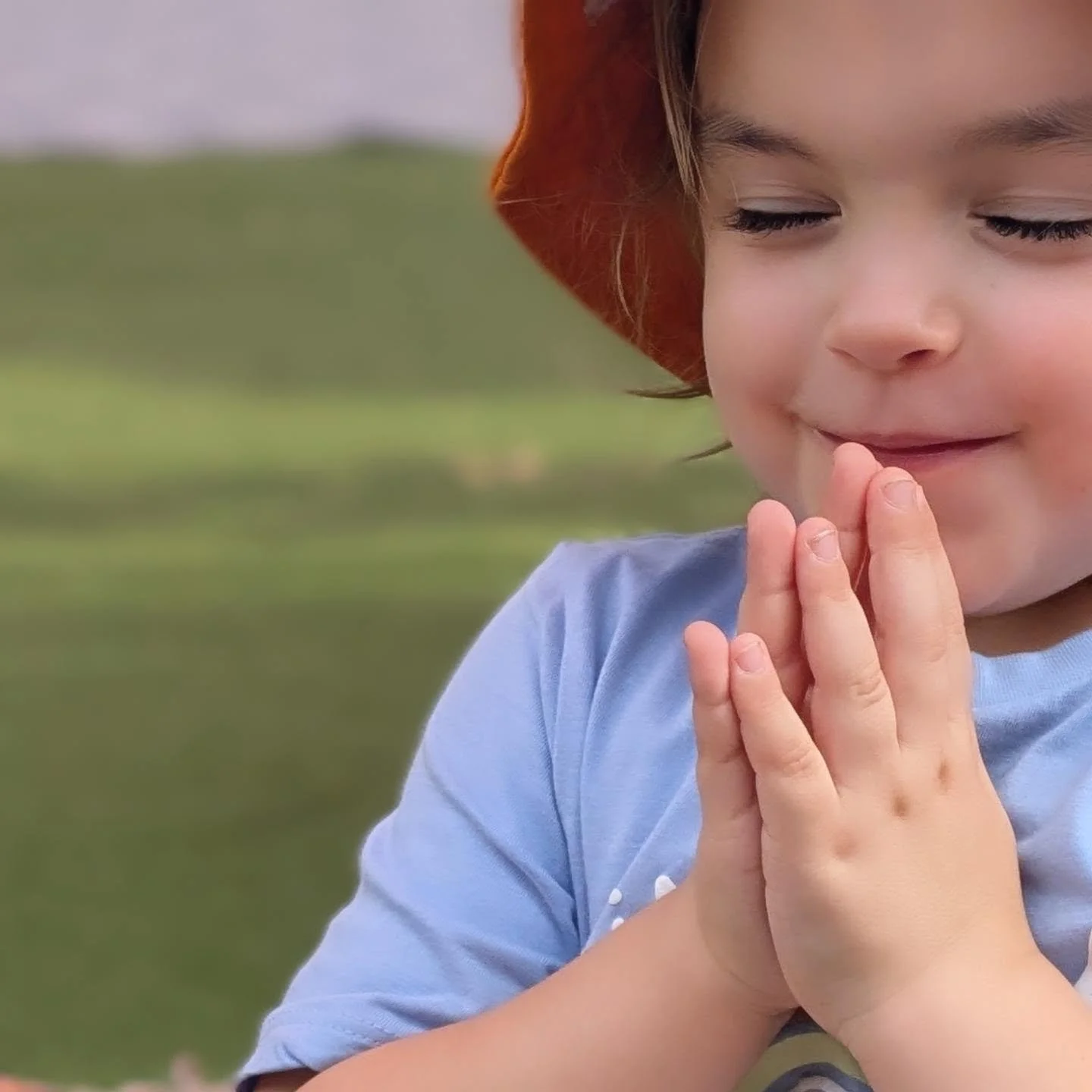 A young child with closed eyes and a slight smile, wearing a red hat and a light blue shirt, clasping their hands together in a peaceful outdoor setting with a grassy background.
