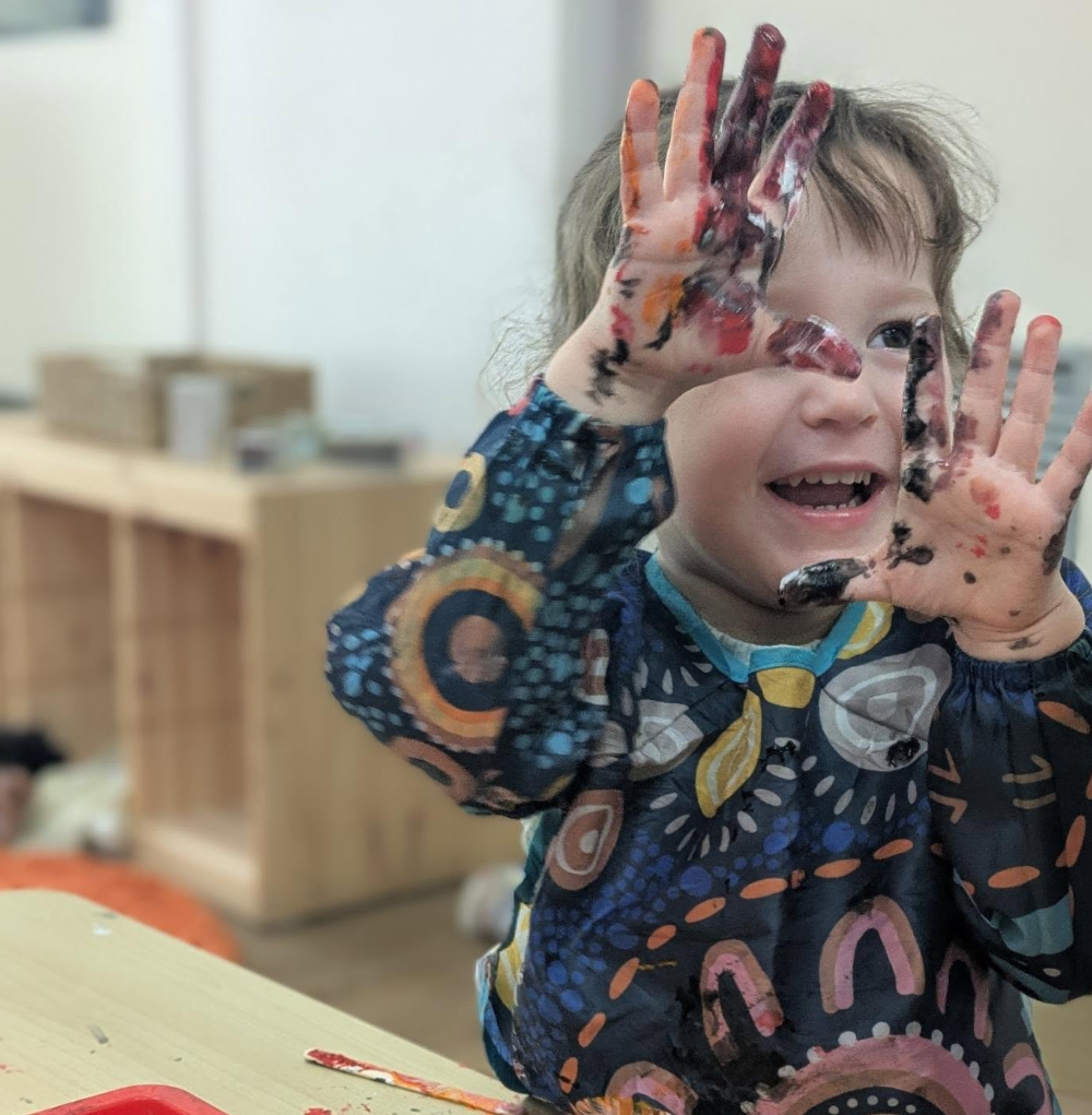 A young child with painted hands smiling and playing, with hair messy and in an indoor setting.