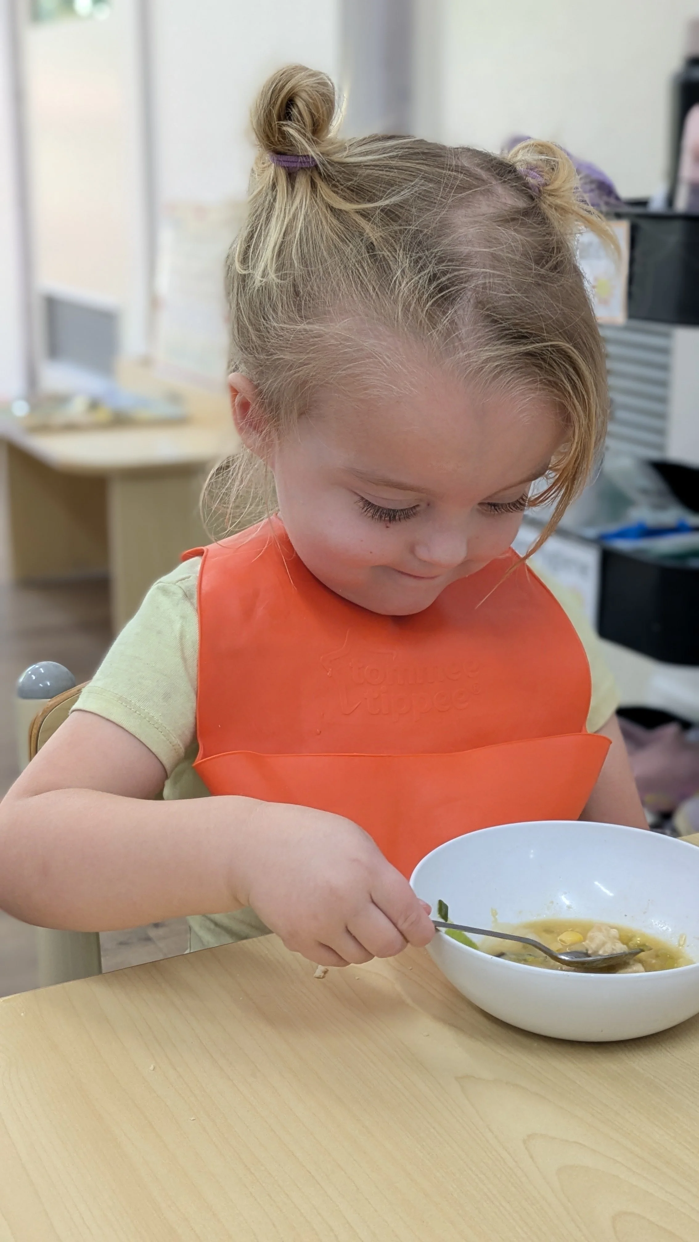 A young girl with blonde hair styled in two buns, wearing an orange bib, eating soup from a white bowl in a classroom setting.