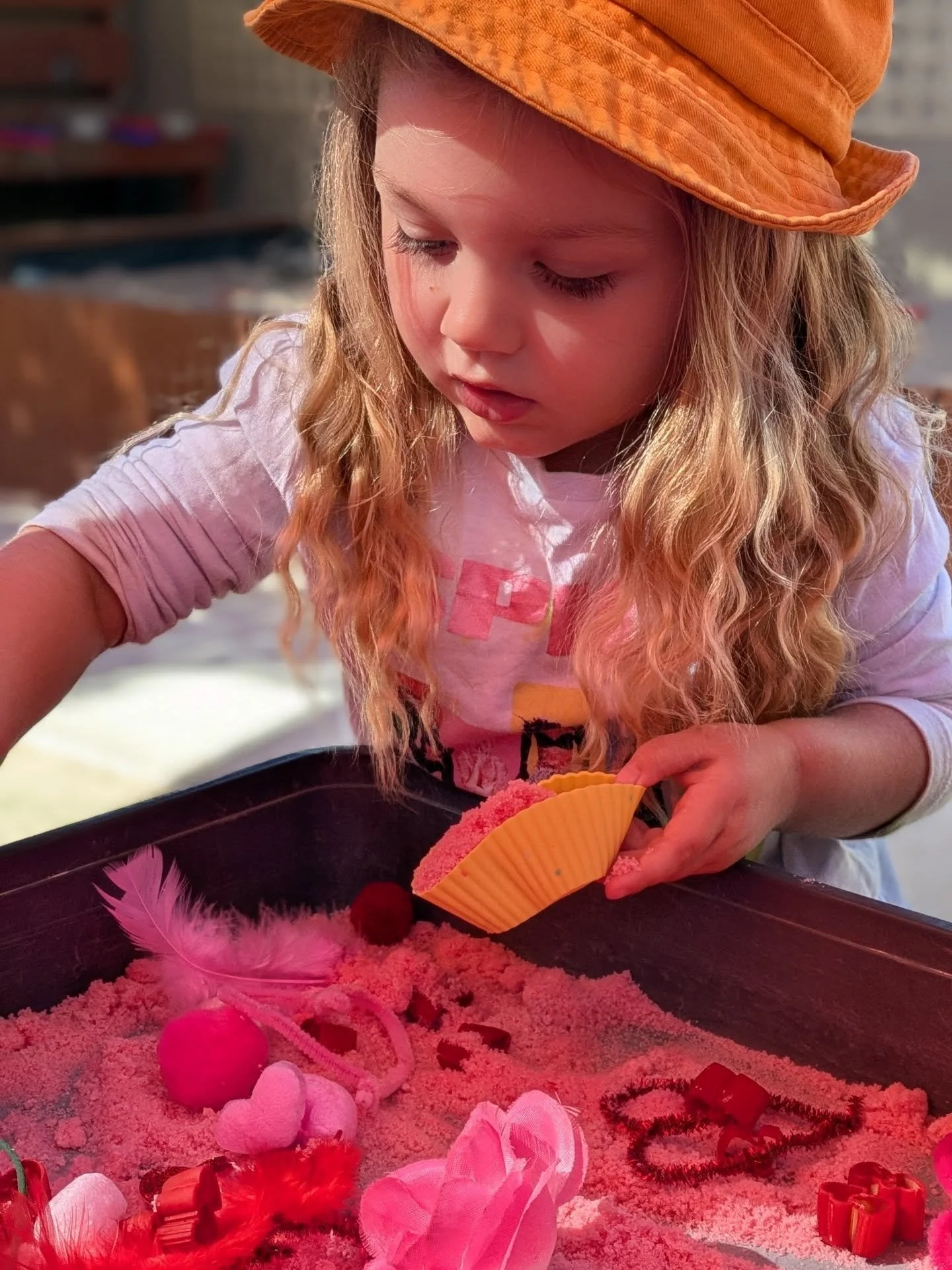 A young girl with long, wavy blonde hair wearing an orange hat, pink shirt, and white sleeves, playing with pink and red Valentine's Day decorations on a table. Brightside Early Learning Centre Child engaging in activity.