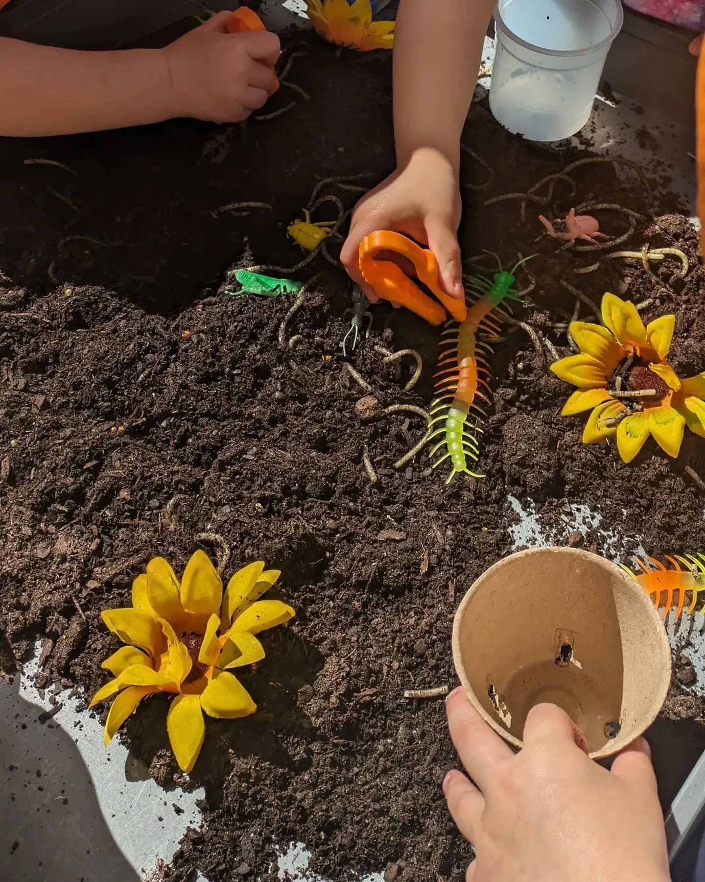 Person planting artificial insects and flowers into dark soil on a garden bed surface.