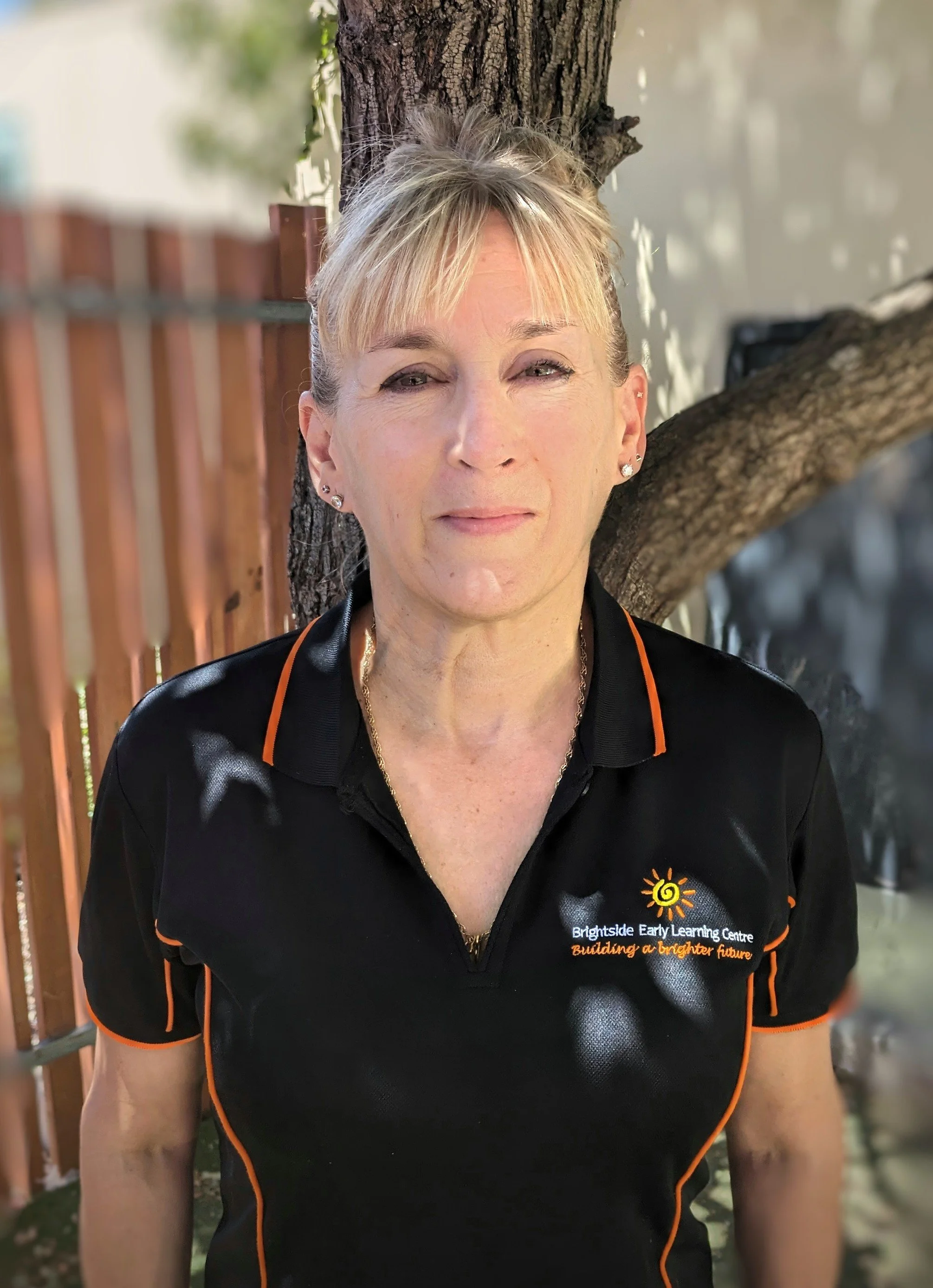 A woman with blonde hair standing outdoors next to a tree. She is wearing a black shirt with orange accents and a logo for Brightside Early Learning Centre.