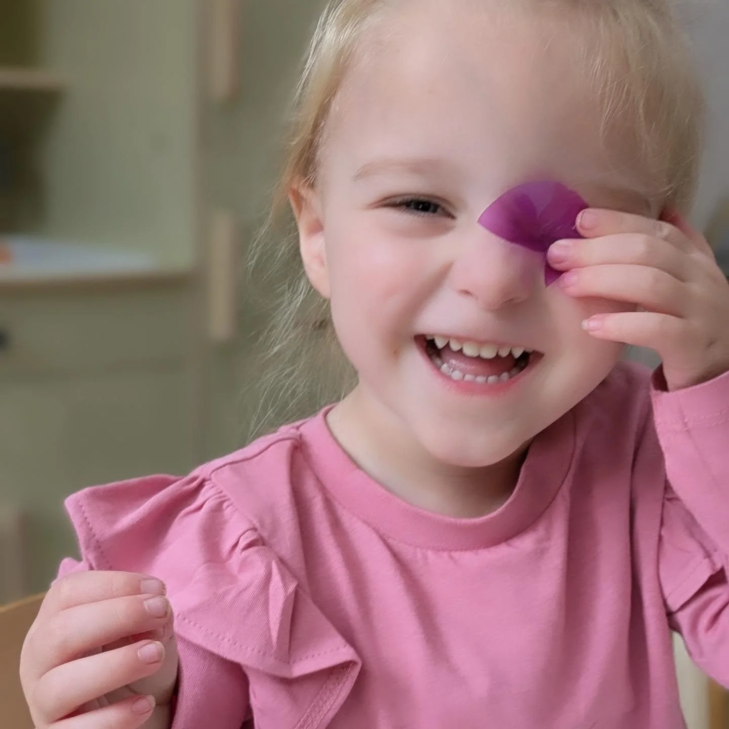 A young girl with blonde hair smiling and holding a purple object to her eye.