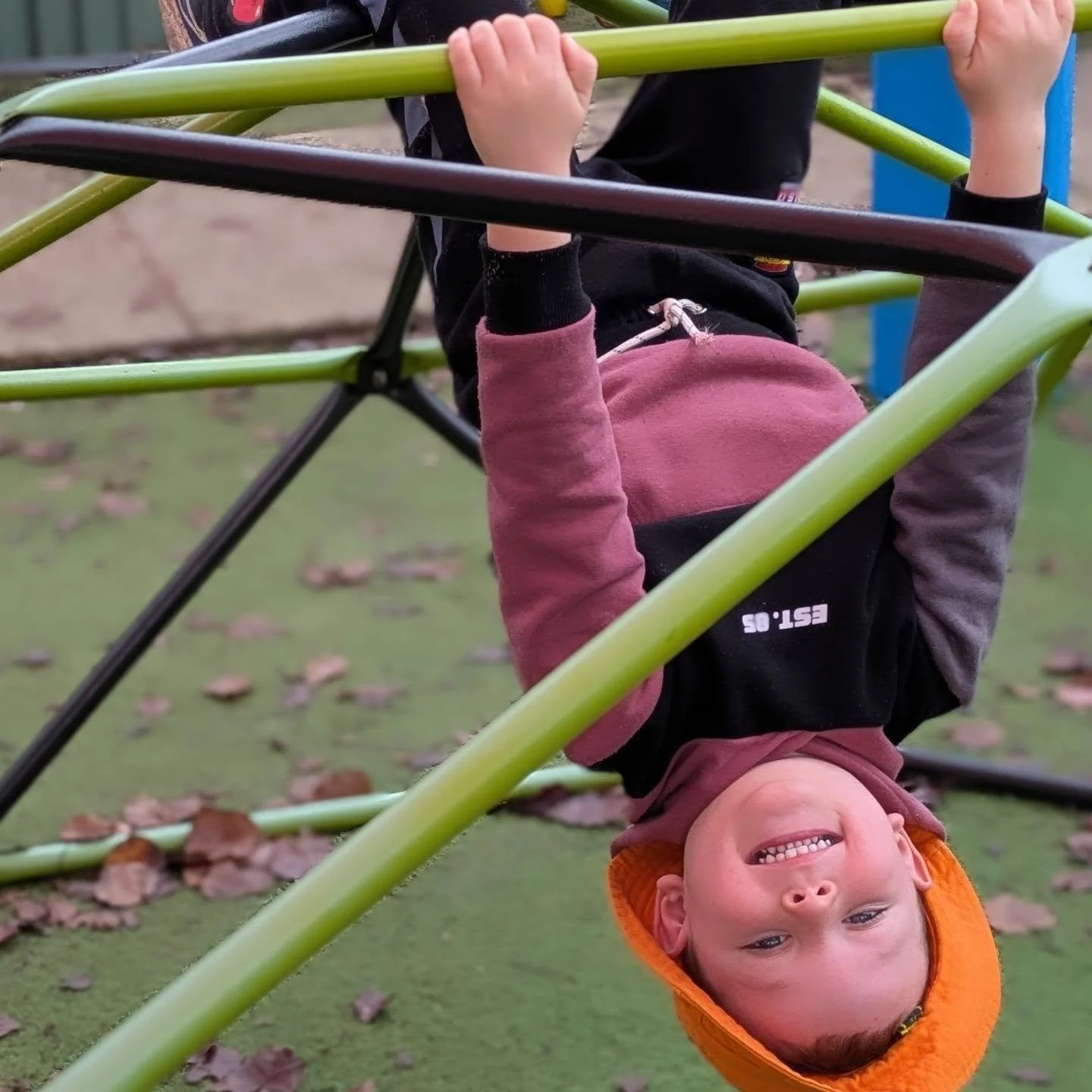 A young boy hanging upside down on a playground climbing structure, smiling and wearing an orange hat and a brown and black jacket.