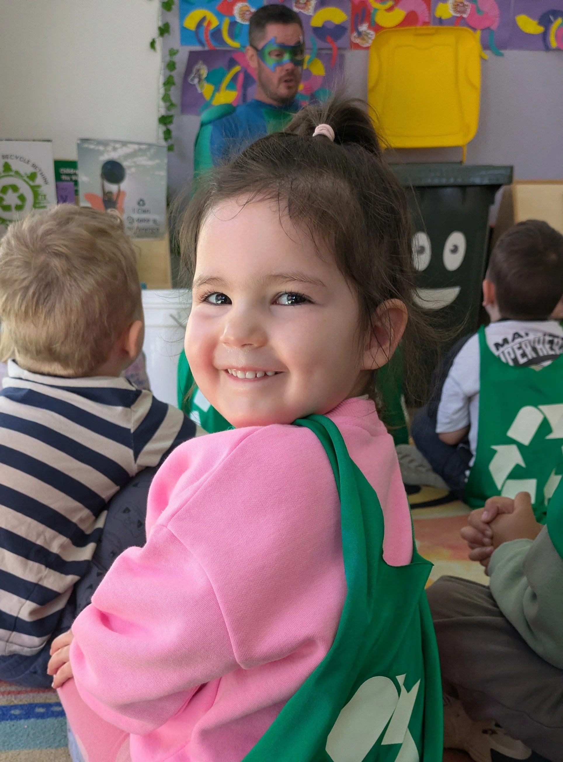 A smiling young girl with brown hair tied in a ponytail held by a pink hairband, wearing a pink hoodie, standing in a classroom with colorful decorations and other children in the background, facing the camera.