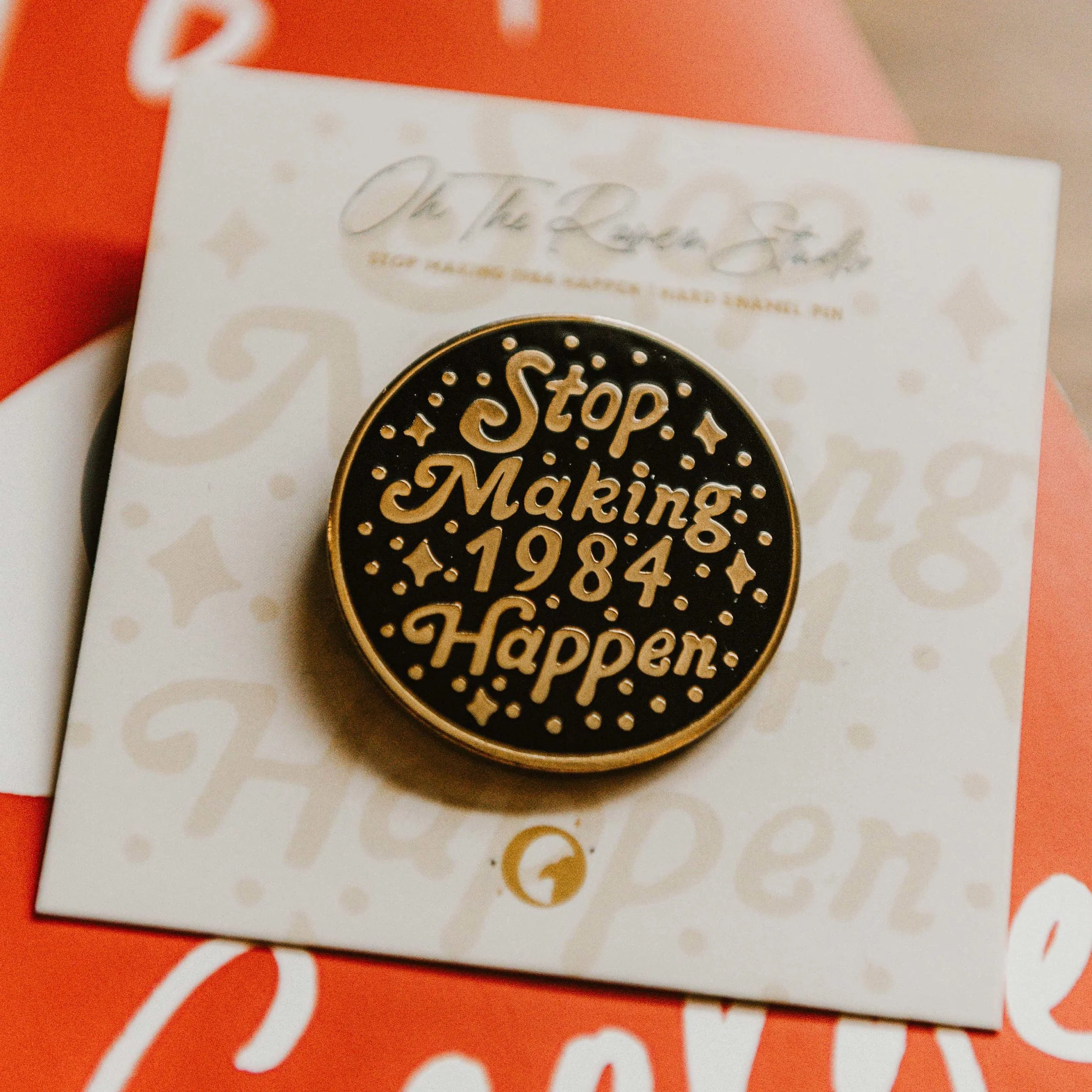Gold and black enamel pin with the quote 'Stop Making 1984 Happen' hand lettered on white backing card with faint decorative text and patterns.
