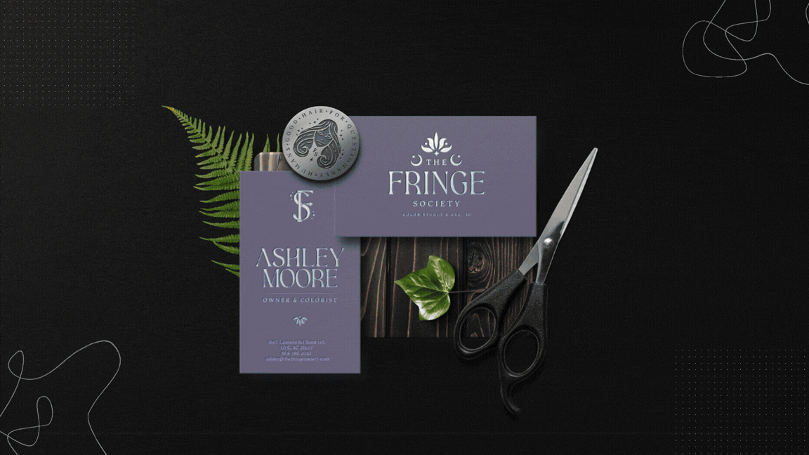 Business cards, a scissors, a fern leaf, and a button with a woman's face are arranged on a black background.