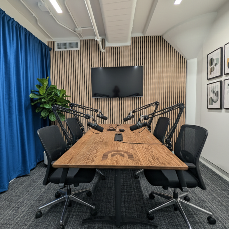 A modern conference room with a wooden table, six black chairs, and six microphones, with an abstract art gallery on the white wall, a large TV screen on a wood-paneled wall, and a blue curtain on the left side.