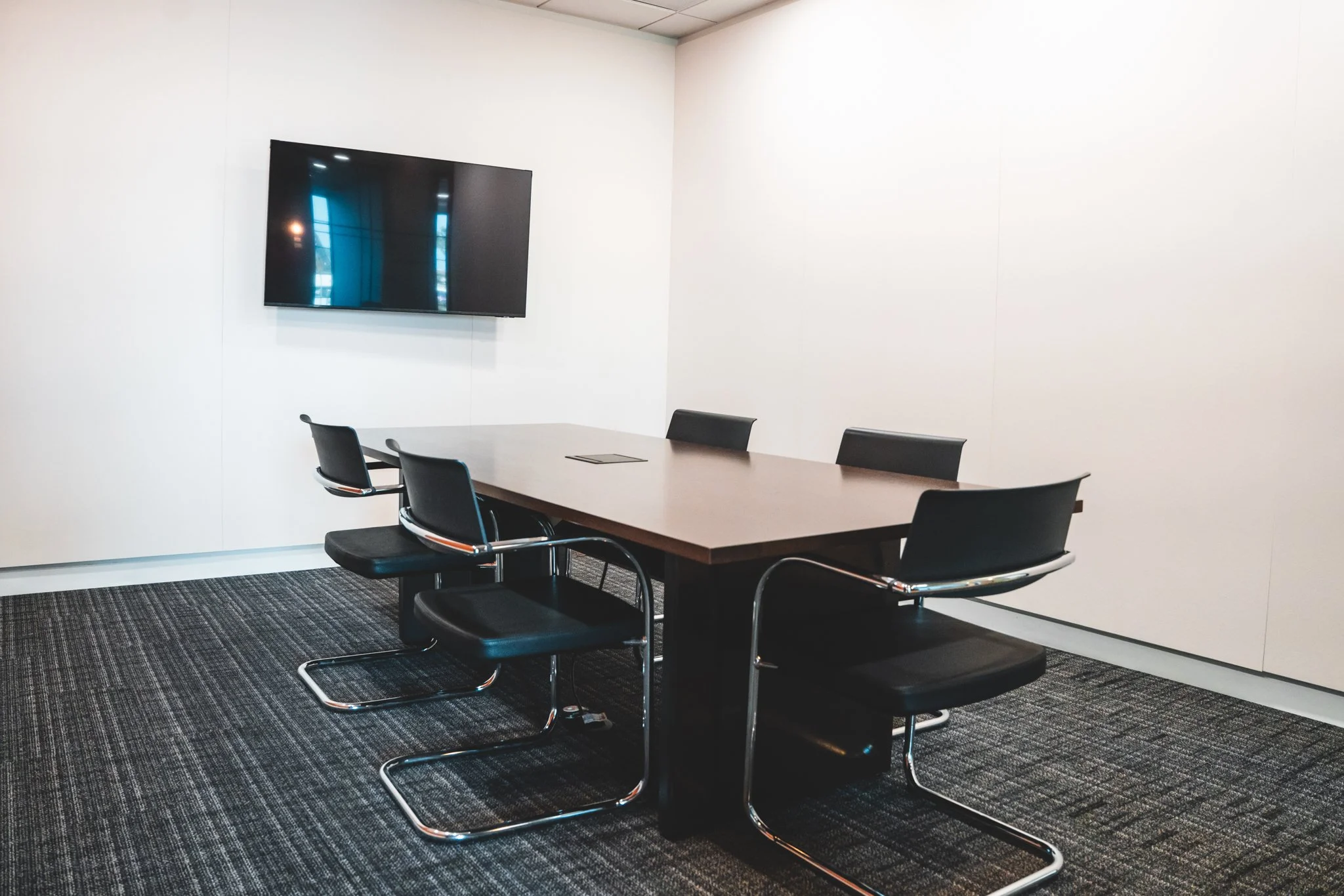 Empty conference room with a long wooden table, four black chairs with chrome frames, a wall-mounted flat-screen TV, and a dark patterned carpet.