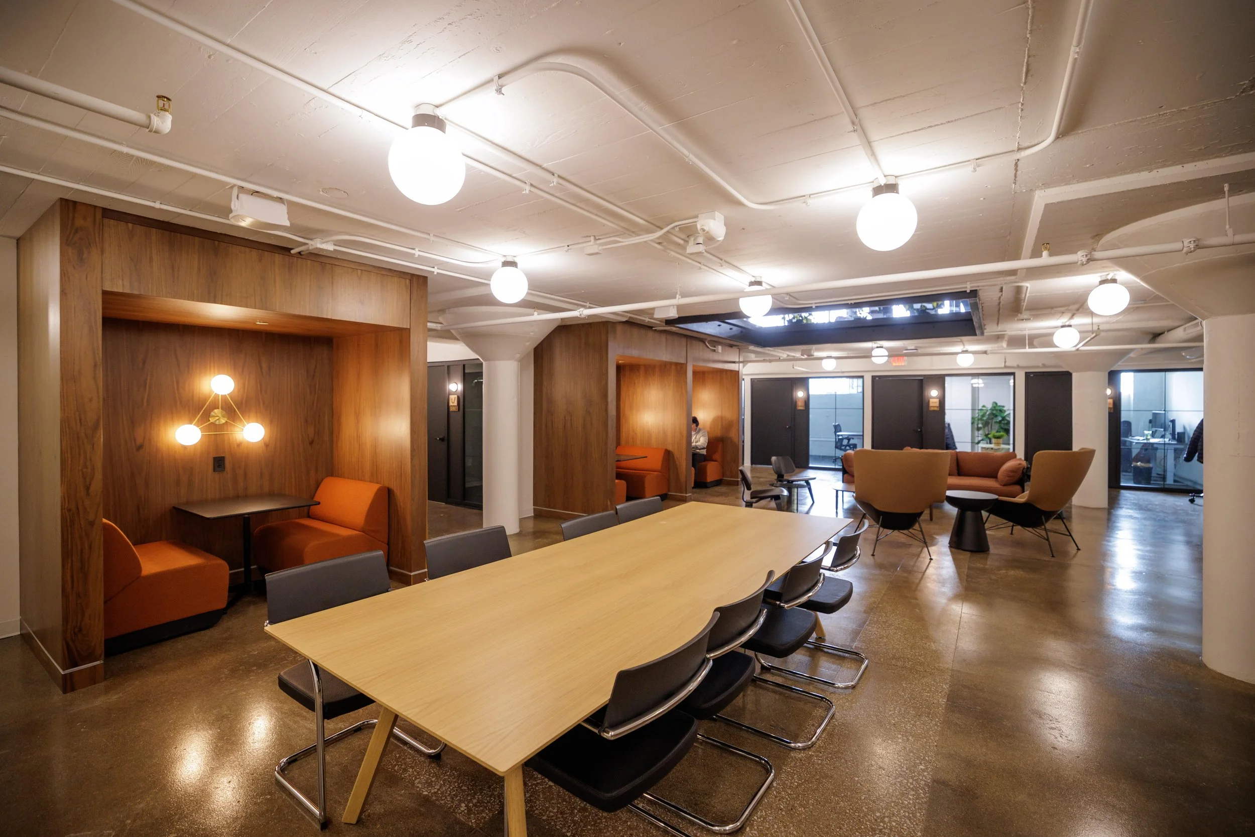 Modern office lounge with a large wooden conference table, black chairs, and various seating areas with sofas and chairs, vases with green plants, and contemporary lighting fixtures.