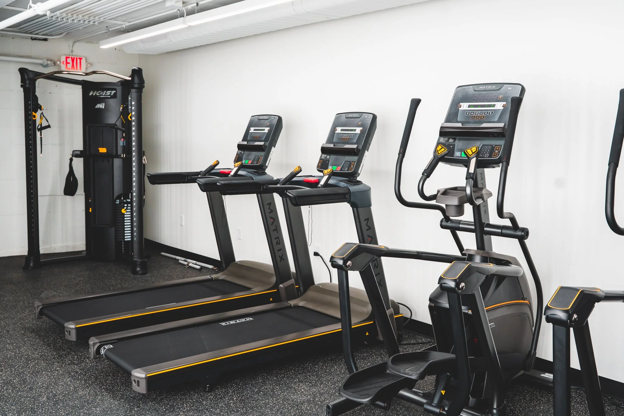 Gym equipment including two treadmills, an elliptical trainer, and a cable machine against a white wall.