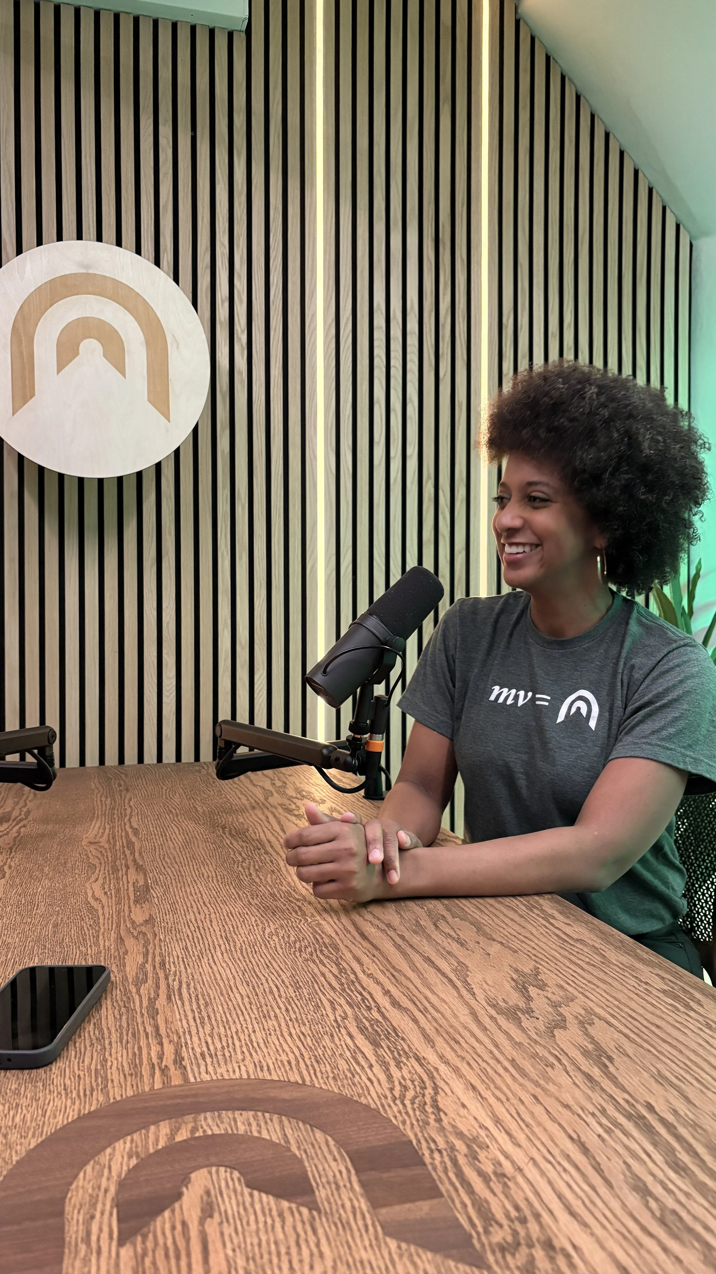 A woman with curly hair smiling and sitting at a wooden table with a microphone in front of her, in a studio with a wooden wall and a logo with concentric arches behind her.