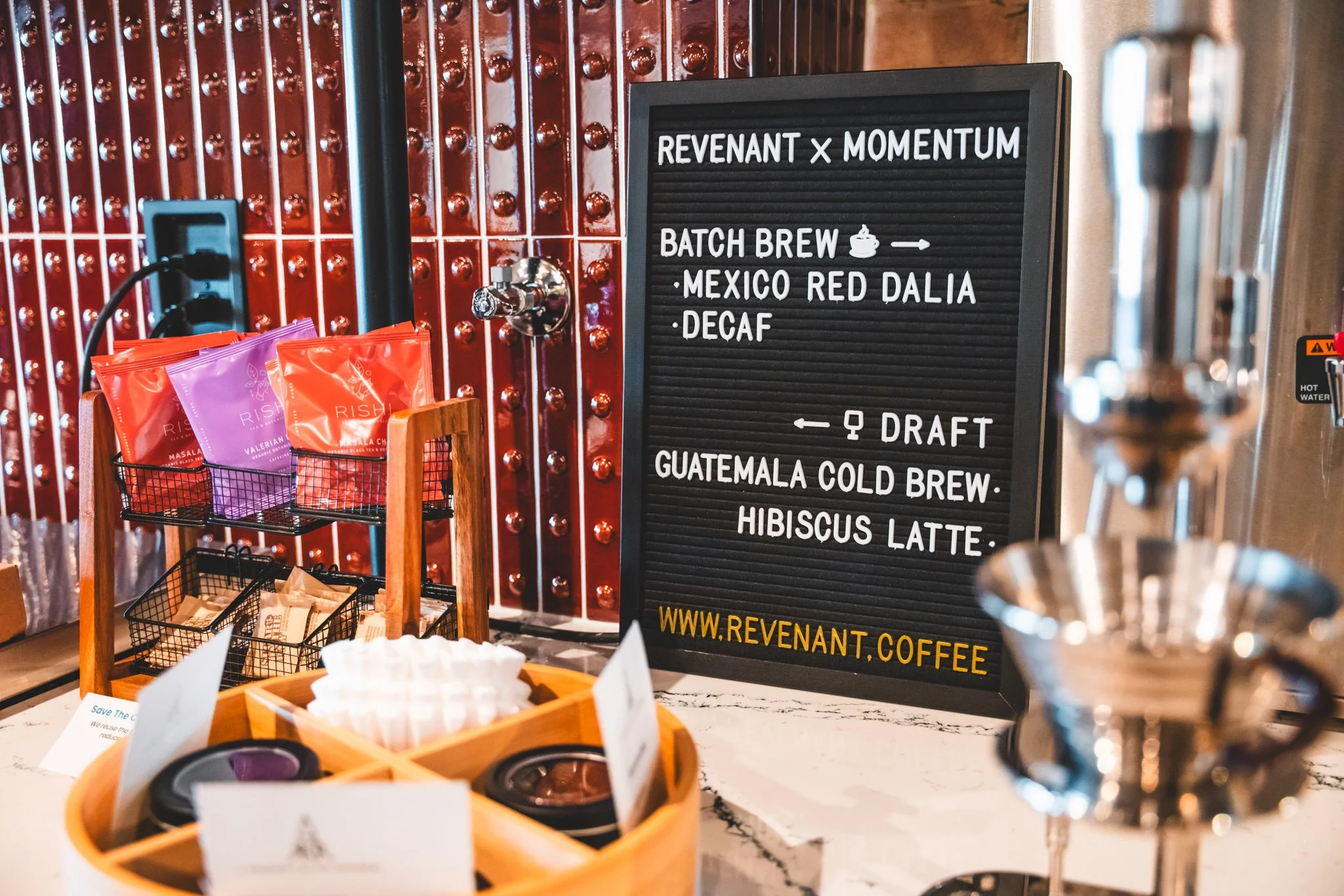 A coffee shop counter displaying various coffee products and a black menu board listing beverages including batch brew, Guatemala cold brew, hibiscus latte, and decaf, with a website URL at the bottom.