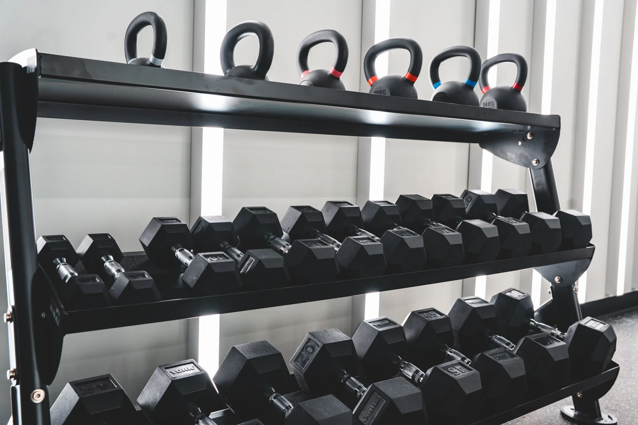 Black dumbbells on three-tier black metal rack, with kettlebells on top, in a gym setting.