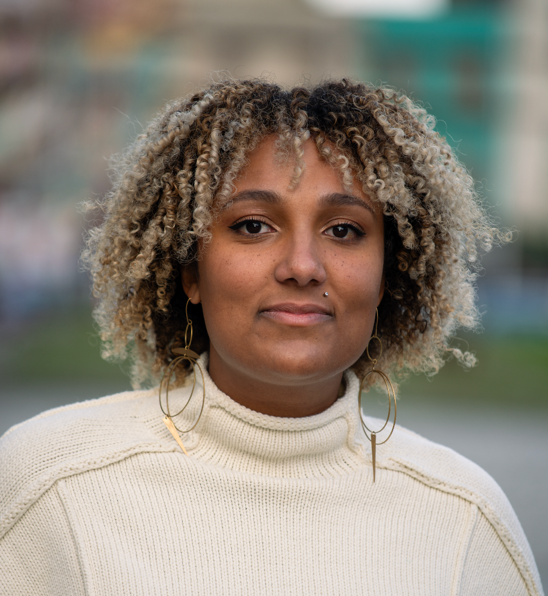 A photo of Tesiah Coleman from the chest up. She is a black woman with coiled tan and black hair to her shoulders, who wears a white sweater and gold hoop earrings. The background of the photo is blurry.