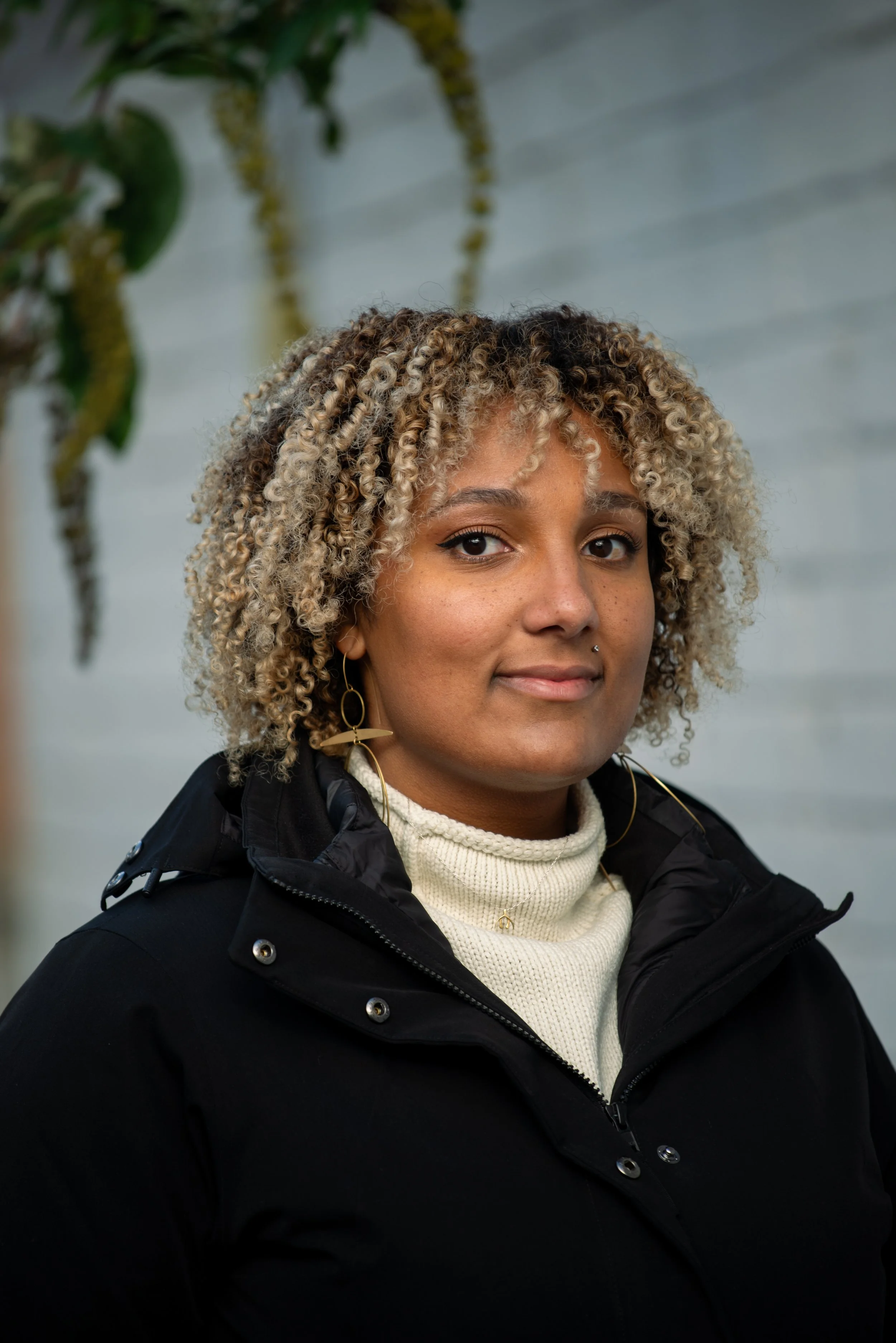 Portrait of a woman with curly blonde and black hair, wearing gold earrings, a beige turtleneck sweater, and a black jacket, standing outdoors with blurred background of a light-colored wall and greenery.