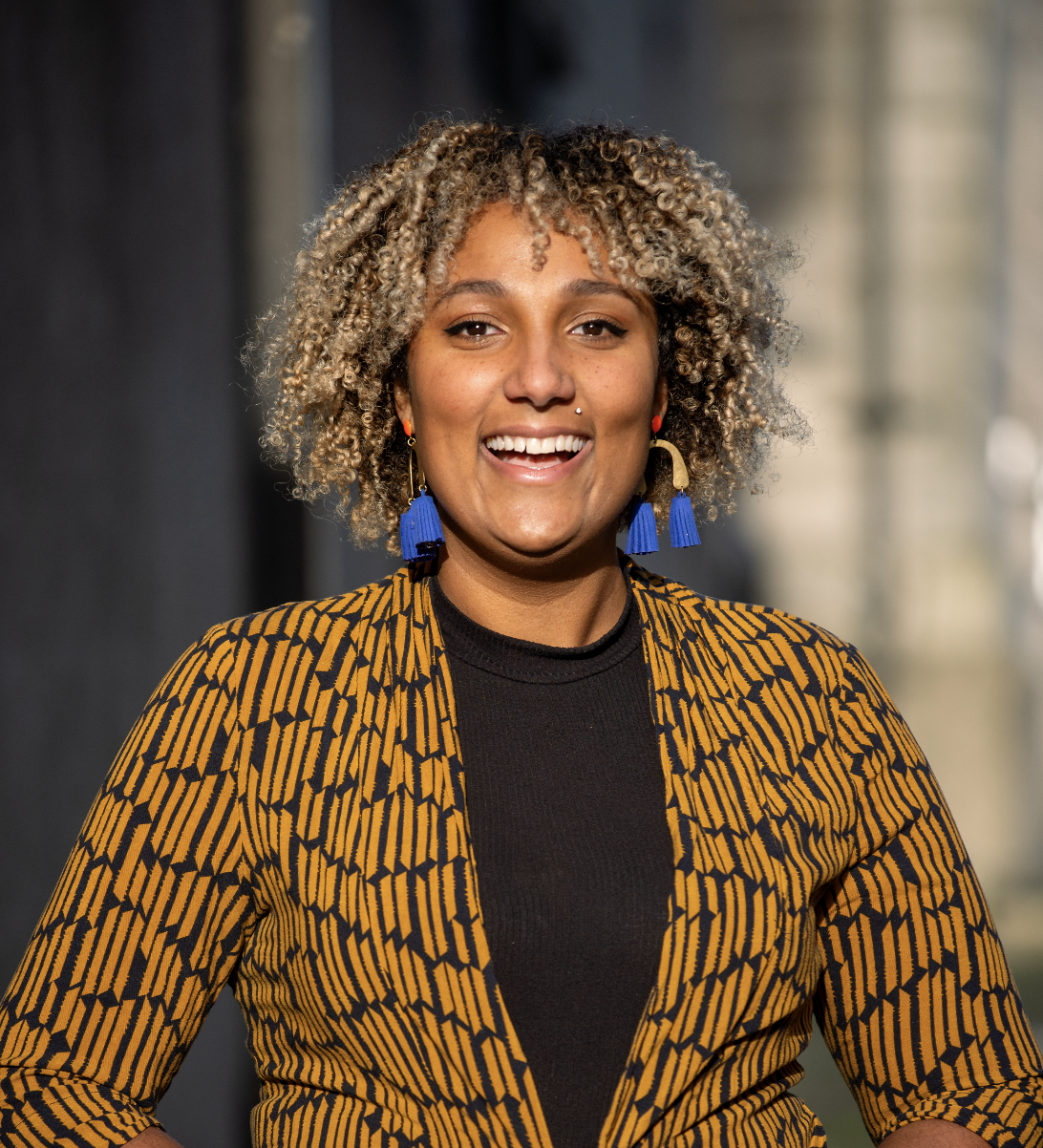 A photo of Tesiah from the waist up. She is a Black woman with coiled tan and black hair, blue earrings, and a yellow and black suit-jacket over a black shirt.