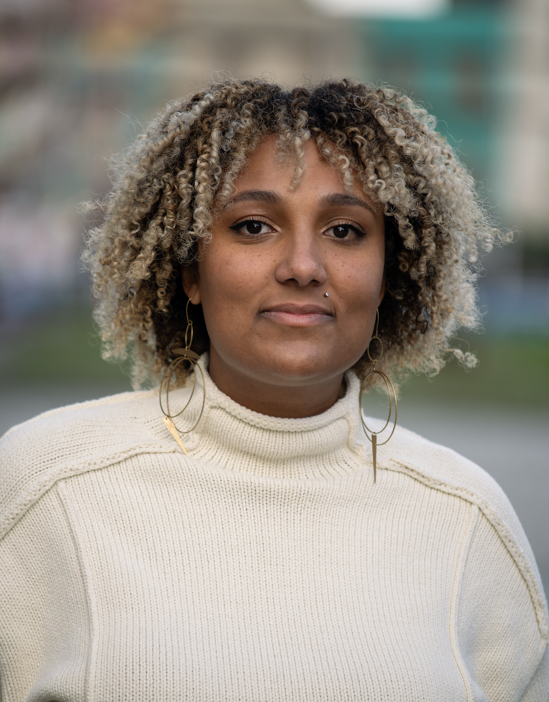 A photo of Tesiah Coleman from the chest up. She is a black woman with coiled tan and black hair to her shoulders, who wears a white sweater and gold hoop earrings. The background of the photo is blurry.