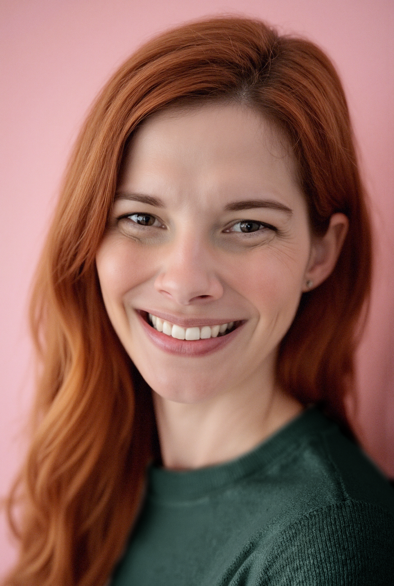 A headshot of a white woman with long, straight red hair in a green sweater against a light pink background. She is looking directly into the camera and smiling.
