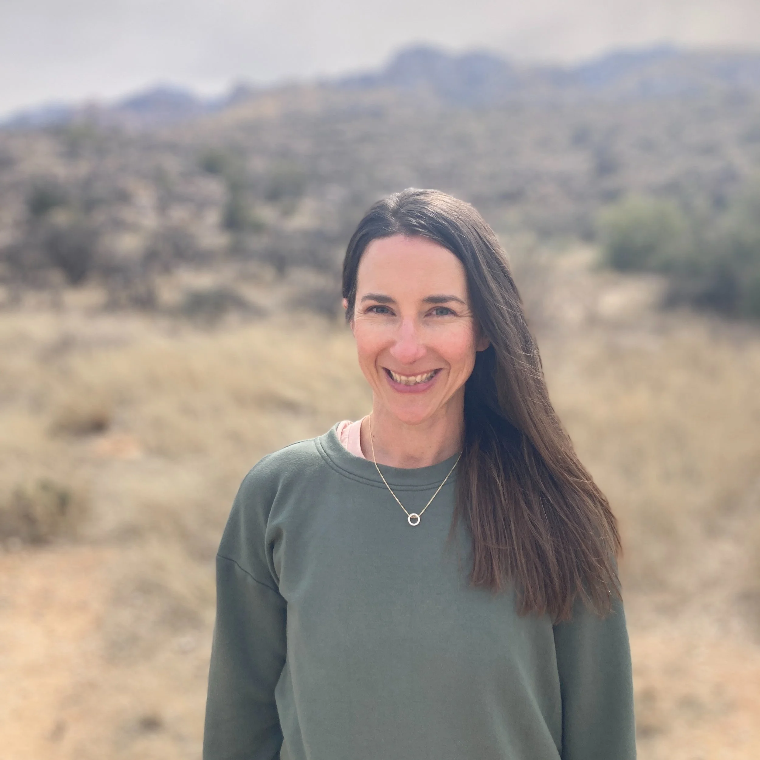 A smiling woman with long brown hair standing outdoors in a desert-like area with hills in the background.