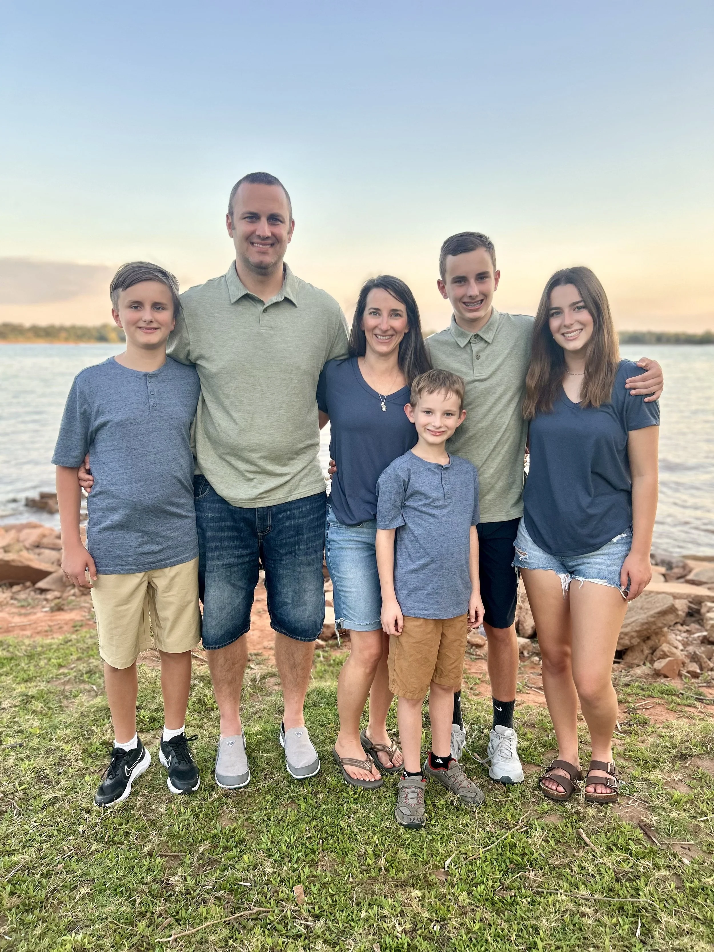 A family of seven standing outdoors near a body of water during sunset, with three children, two teenagers, and two adults, smiling and posing for the photo.