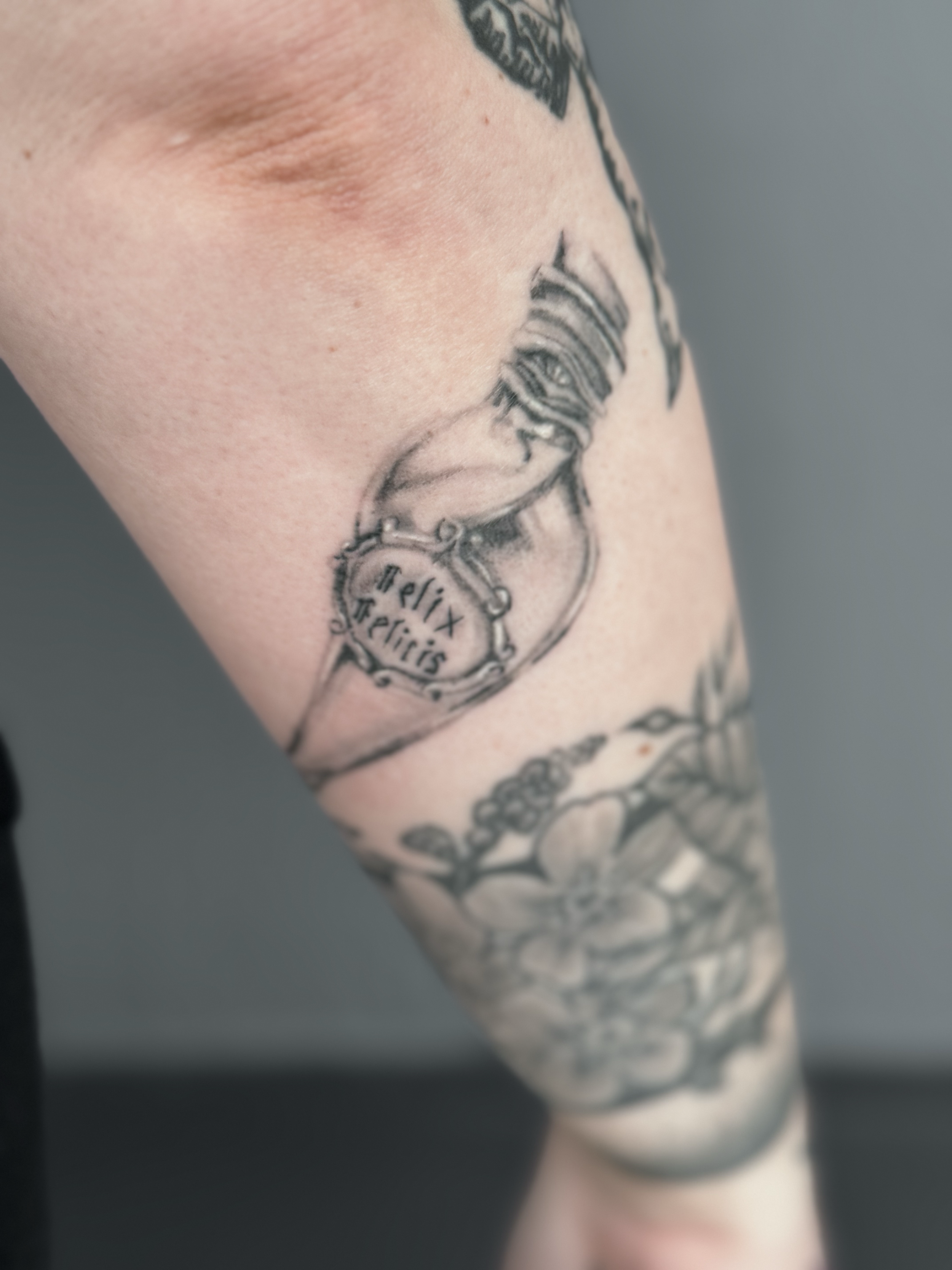 Tattoo on person's upper arm depicting a pocket watch with the text ' Felix Felix' on the clock face, surrounded by decorative elements.