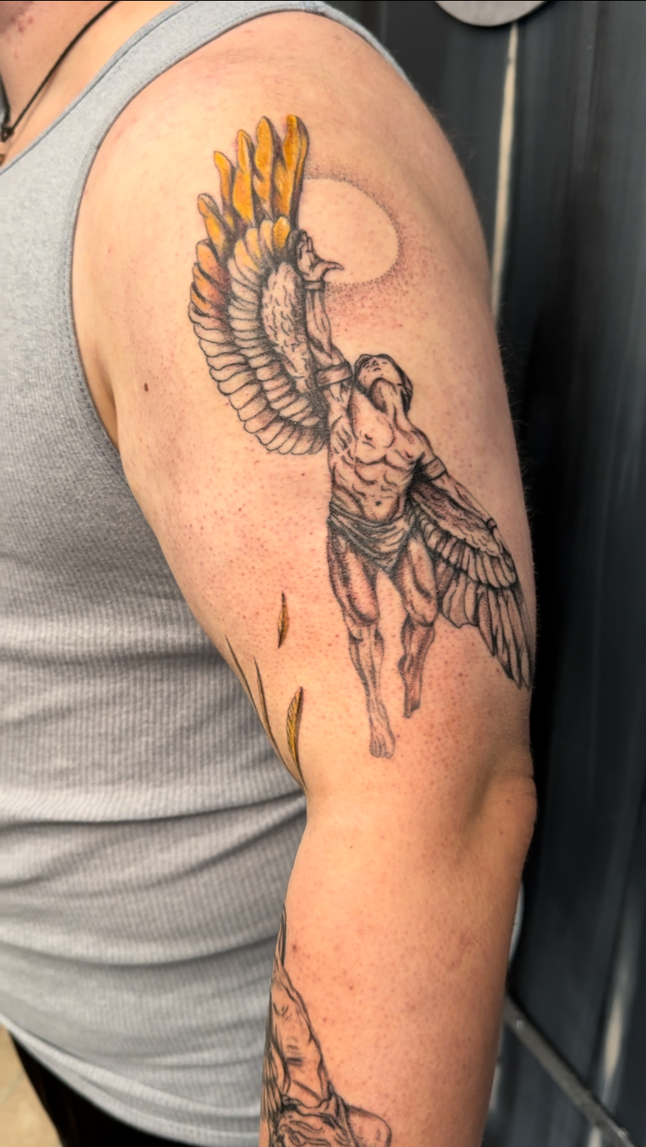 A tattoo of a muscular man with wings flying up, holding a tall staff with a bird's head on top, with a full moon in the background and some falling leaves.