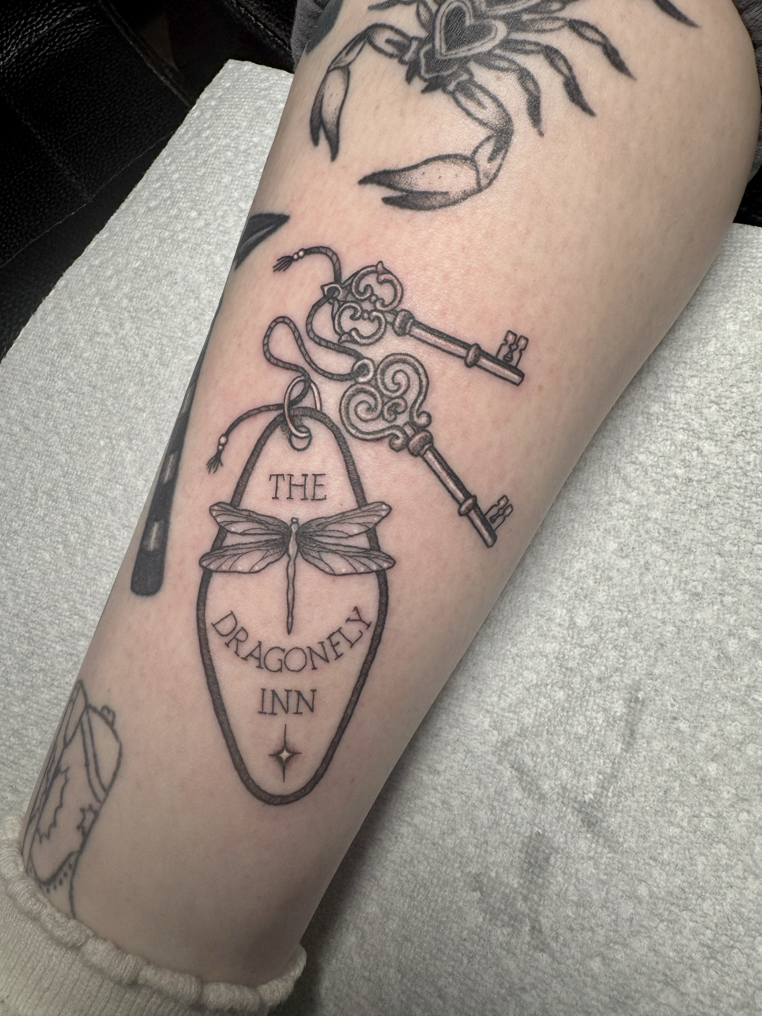 Tattoo of a vintage keyring with multiple keys, a butterfly in a frame with the words "The Dragonfly Inn," and a small star underneath.