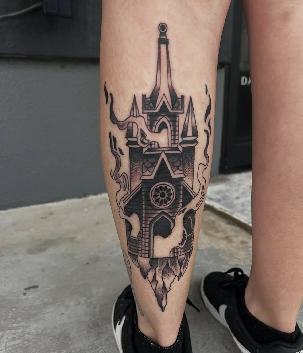 Black and gray tattoo of a haunted house with swirling lines on a person's calf.