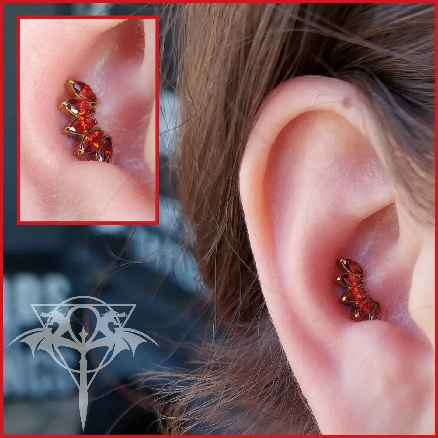A close-up of a person's ear with a red gemstone cartilage jewelry piece in a curved design. An inset shows a detailed view of the jewelry, which features multiple red gemstones set in gold. There is a metallic logo with bat wings and a circle at the