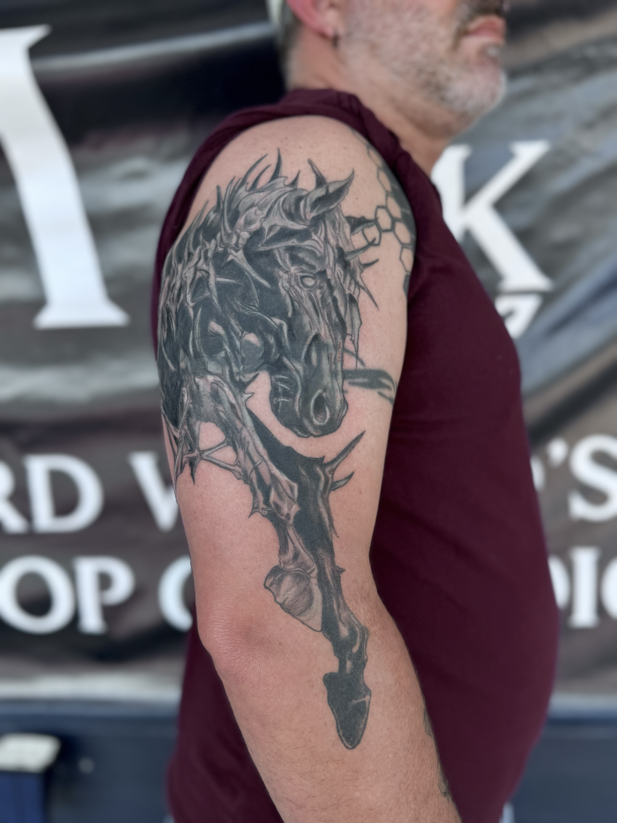 Tattoo of a black horse's head with armor-like details on the upper arm of a man wearing a sleeveless maroon shirt.