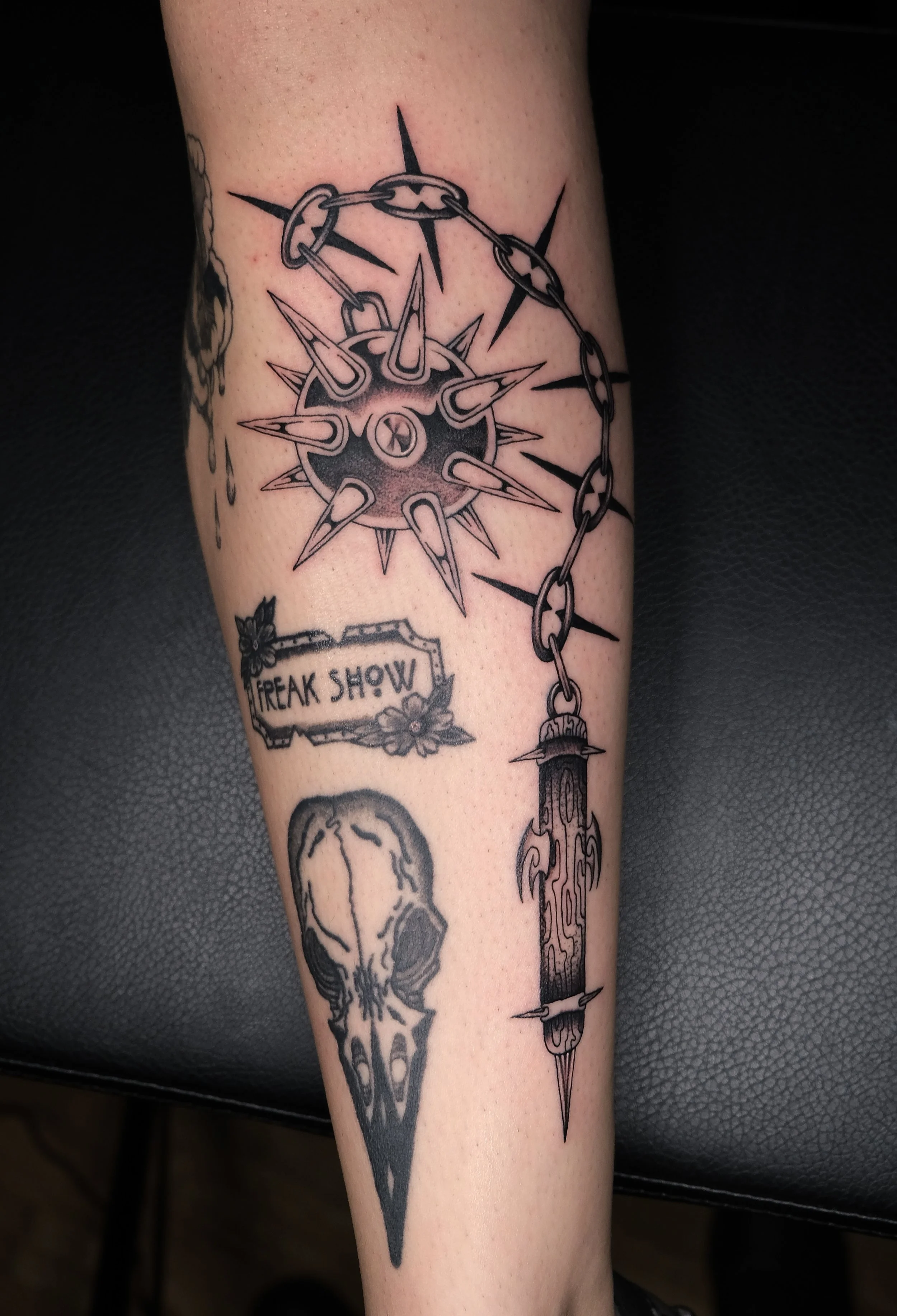 Black and gray tattoo featuring a spiked compass with chains, a skull, a dagger, and a sign that reads 'FREAK SHOW' on a person's arm.