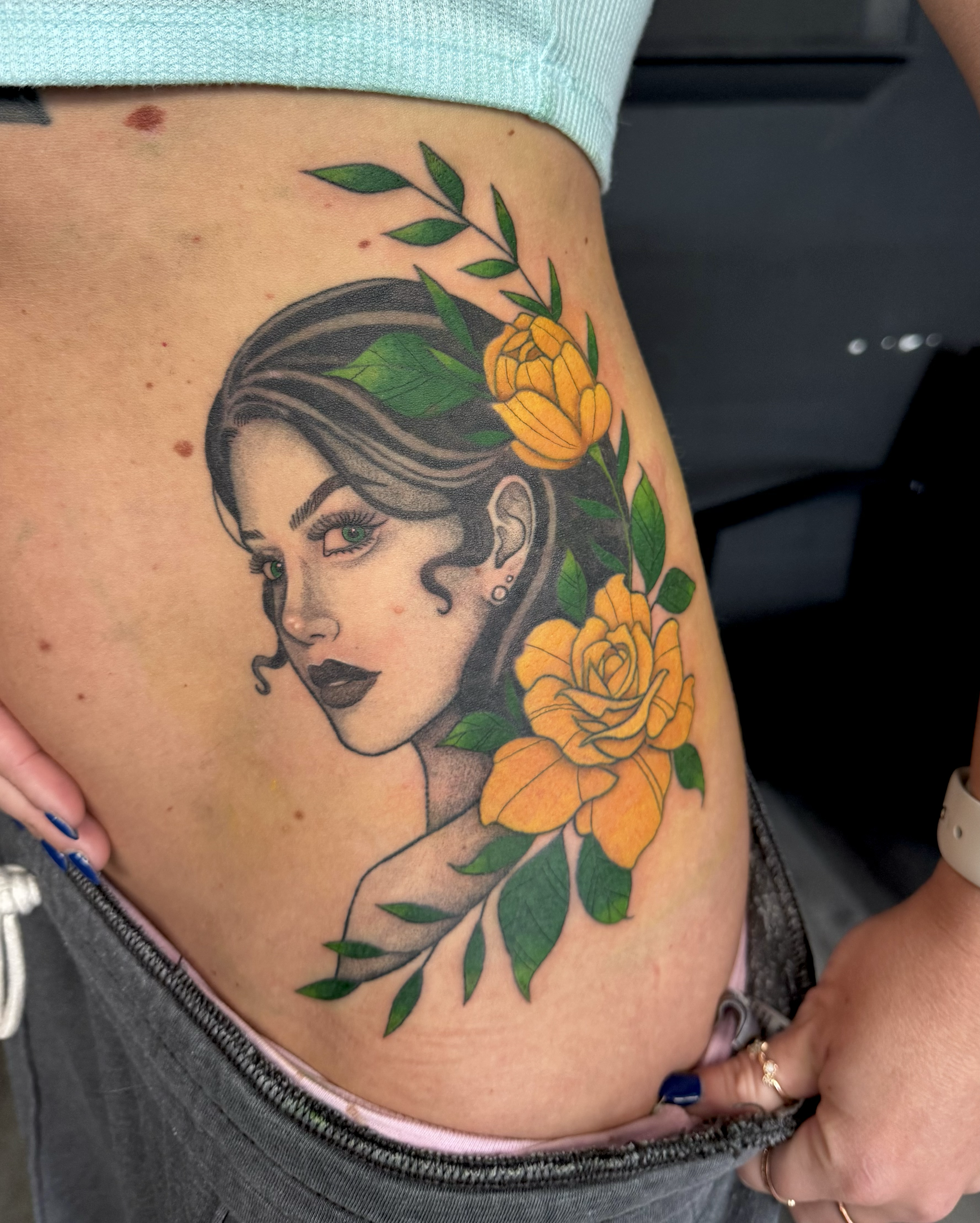 Tattoo of a woman with styled hair, dark lipstick, and earrings, surrounded by yellow roses and green leaves.