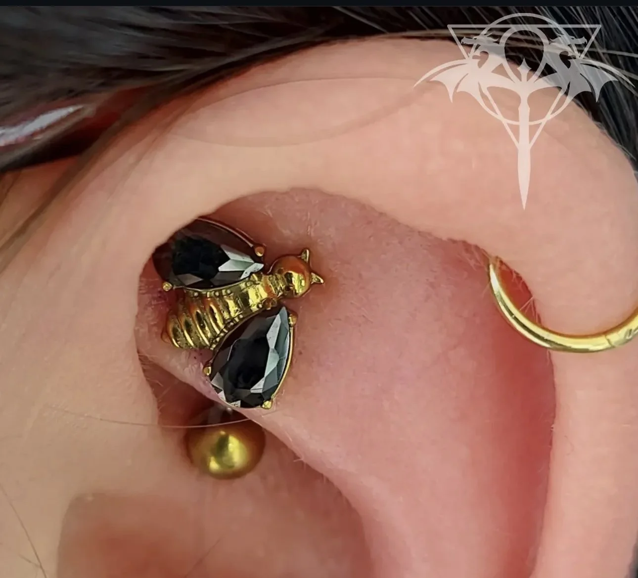 Close-up of a person's ear with multiple earrings, including a gold hoop, a black gemstone stud, and a gold and black gemstone earring, with dark hair partly visible.