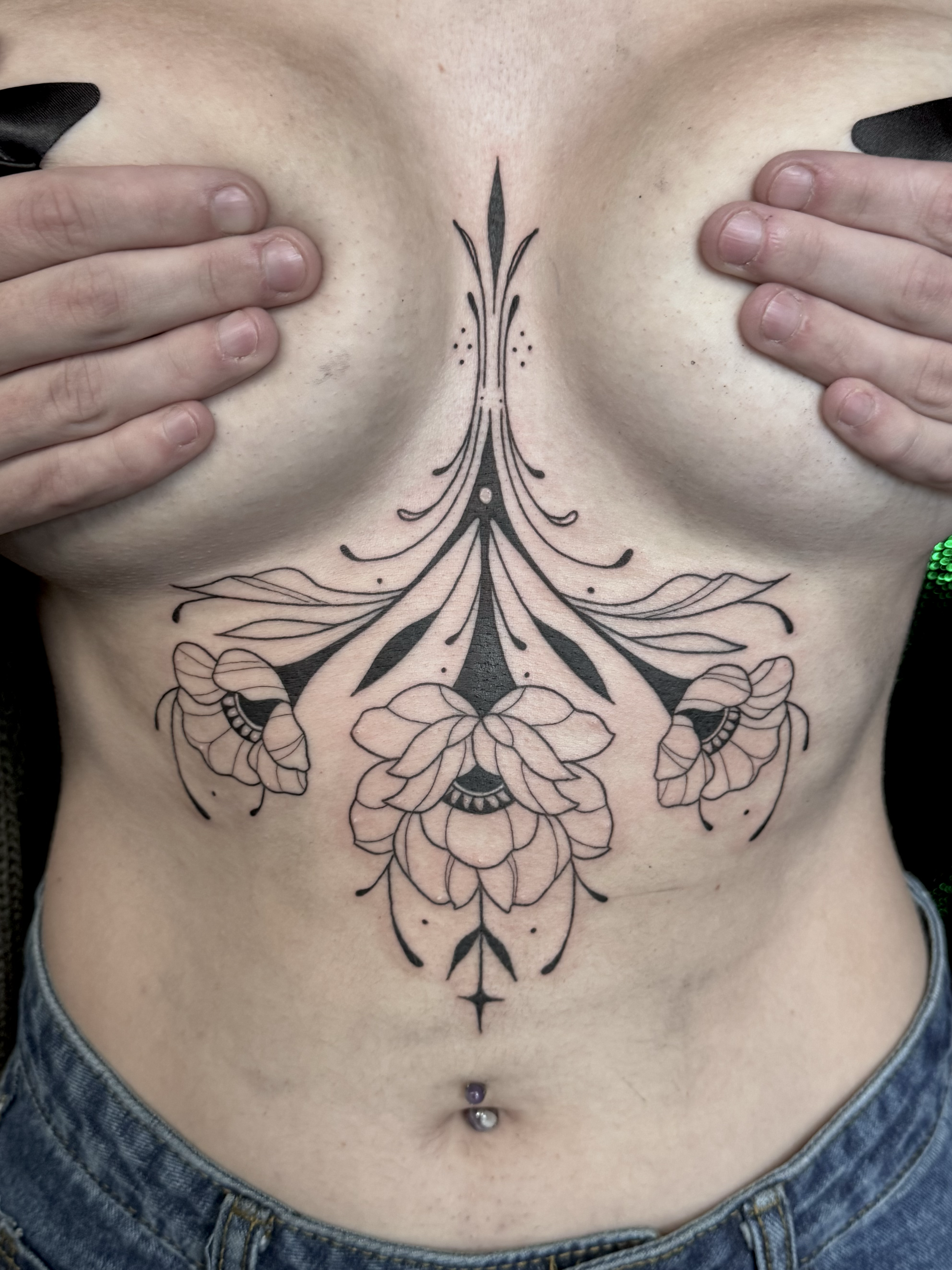 Tattoo of floral and geometric design on person's chest and upper stomach, with hands pressing the breasts to reveal the tattoo, and a navel piercing below.