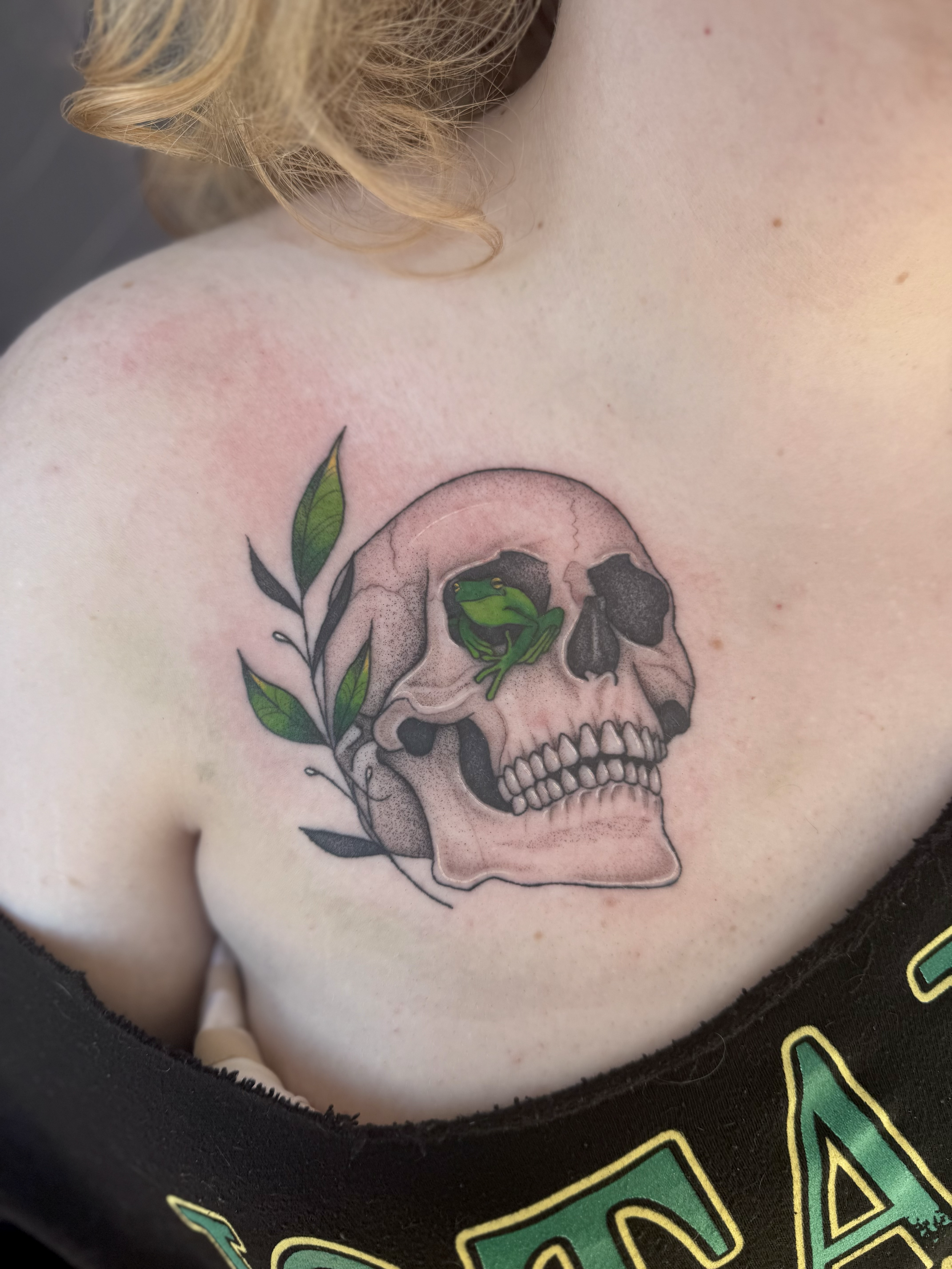 Tattoo of a human skull with green leaves and a small green frog on top of the skull on a person's shoulder.