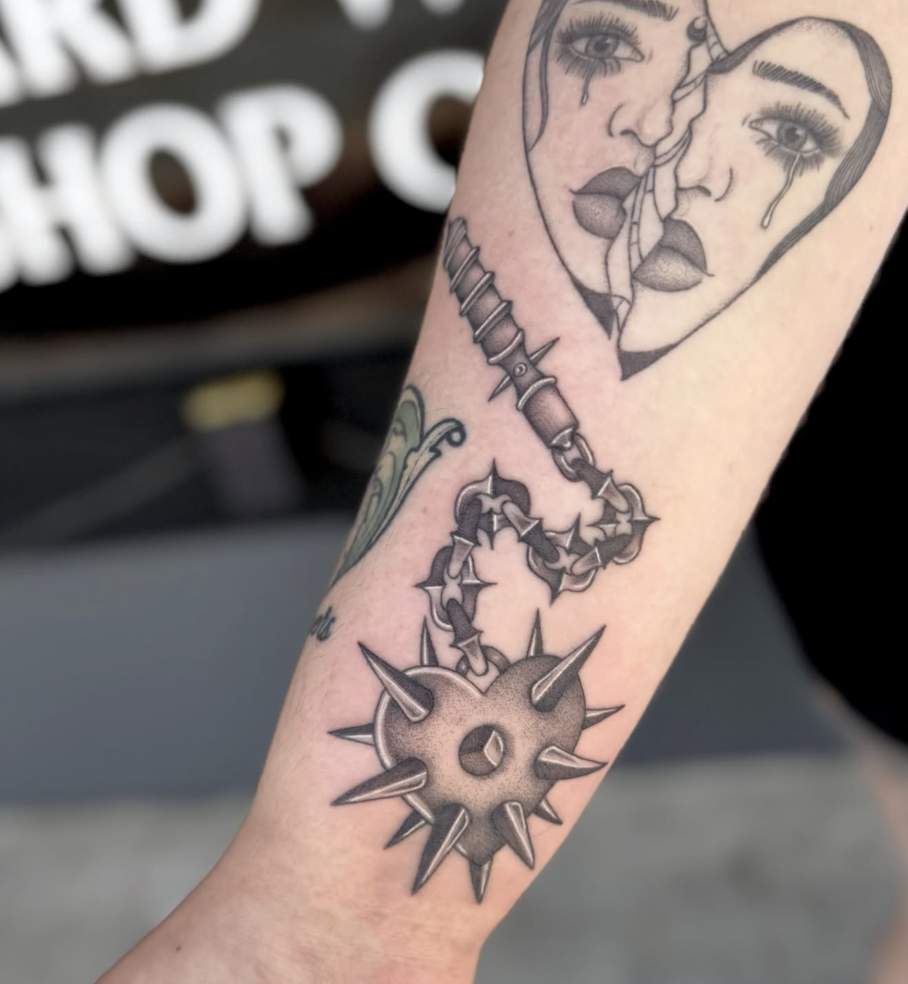 Tattoo of a heart-shaped design with a crack, featuring two faces with tearful eyes near the top and a spiked gear and chain below.