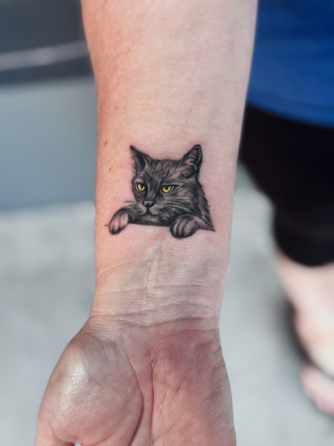 Tattoo of a gray cat with yellow eyes resting its paws on a surface on a person's forearm.