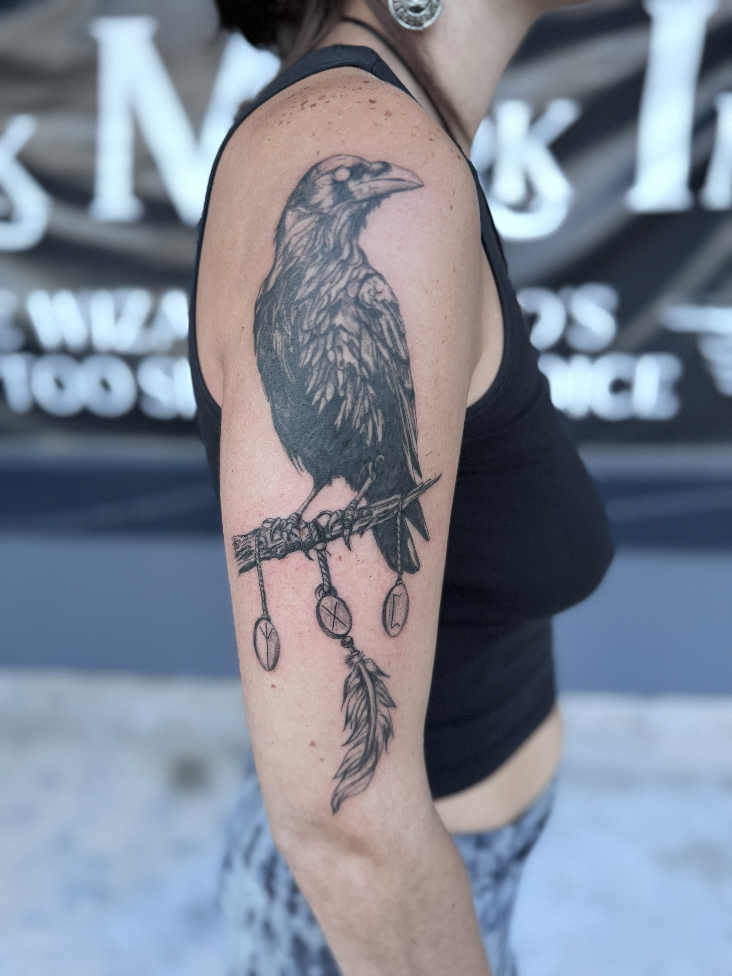 Black tattoo of a raven perched on a branch with feathers and small ornaments hanging from it on a person's upper arm.