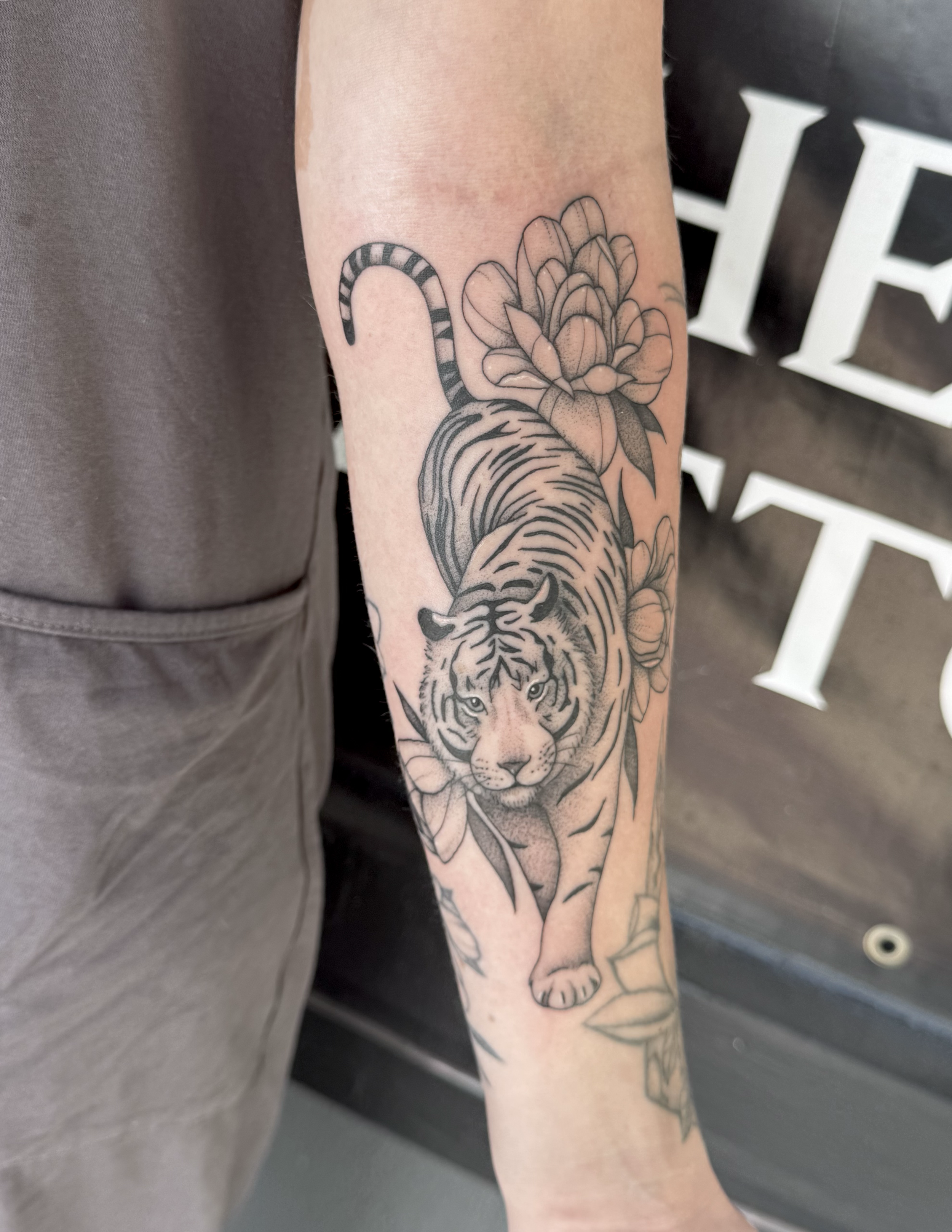 Black and gray tattoo of a tiger walking with flowers on the arm.