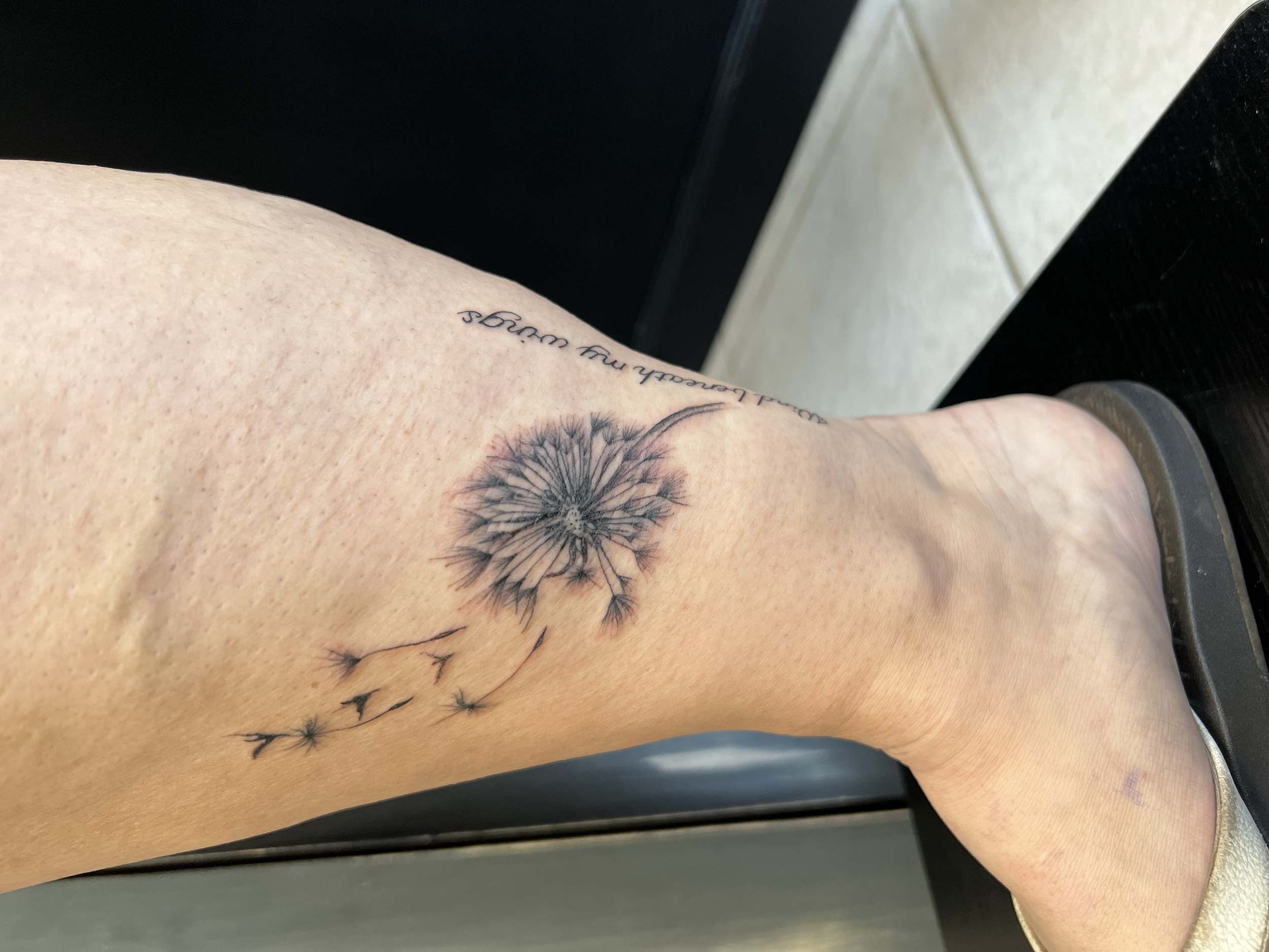 Tattoo of a dandelion flower with its seeds blowing away, on a person's ankle and lower leg.