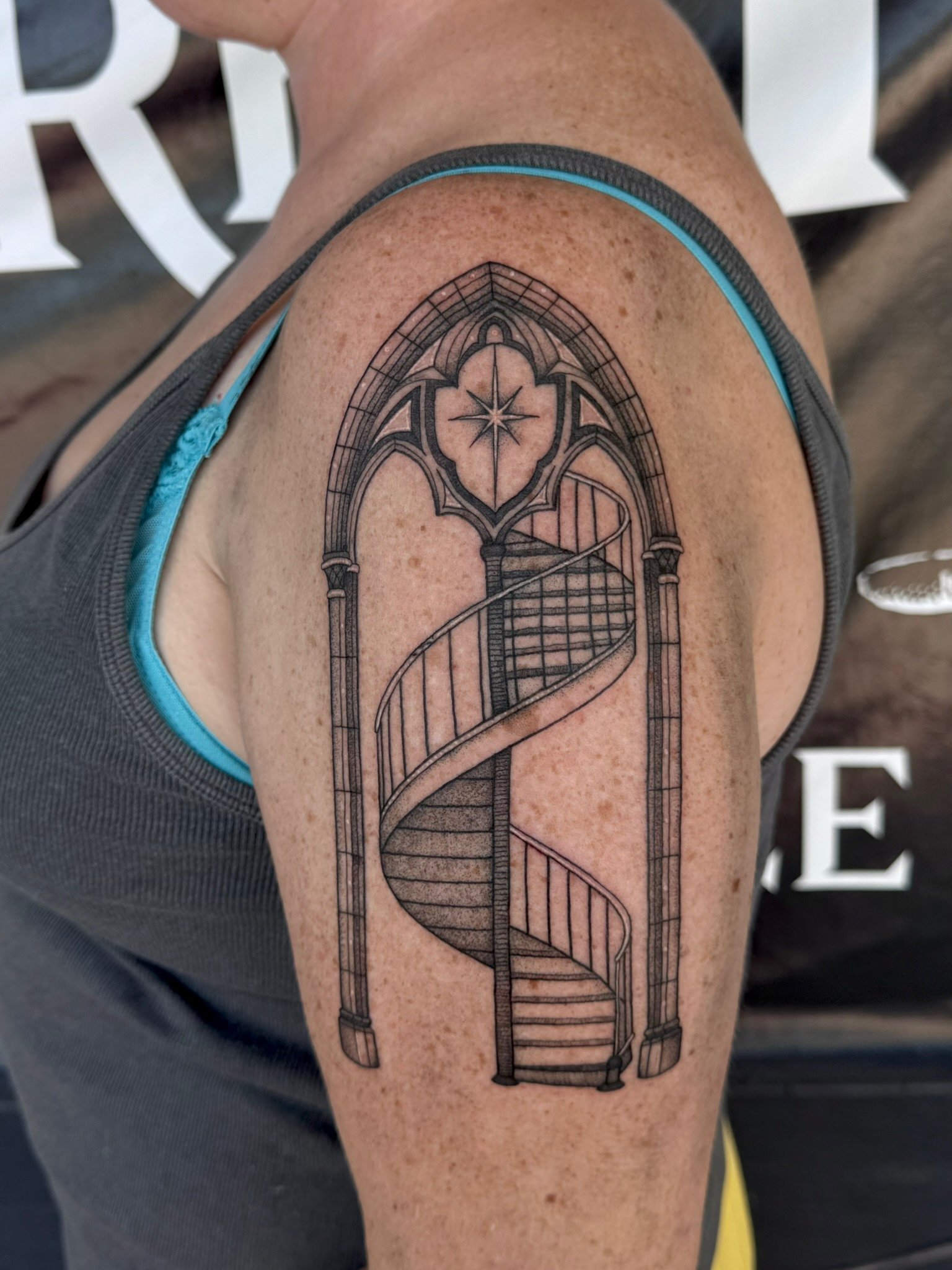Tattoo of a spiral staircase inside a Gothic arch with a star and floral design at the top, on person's upper arm.