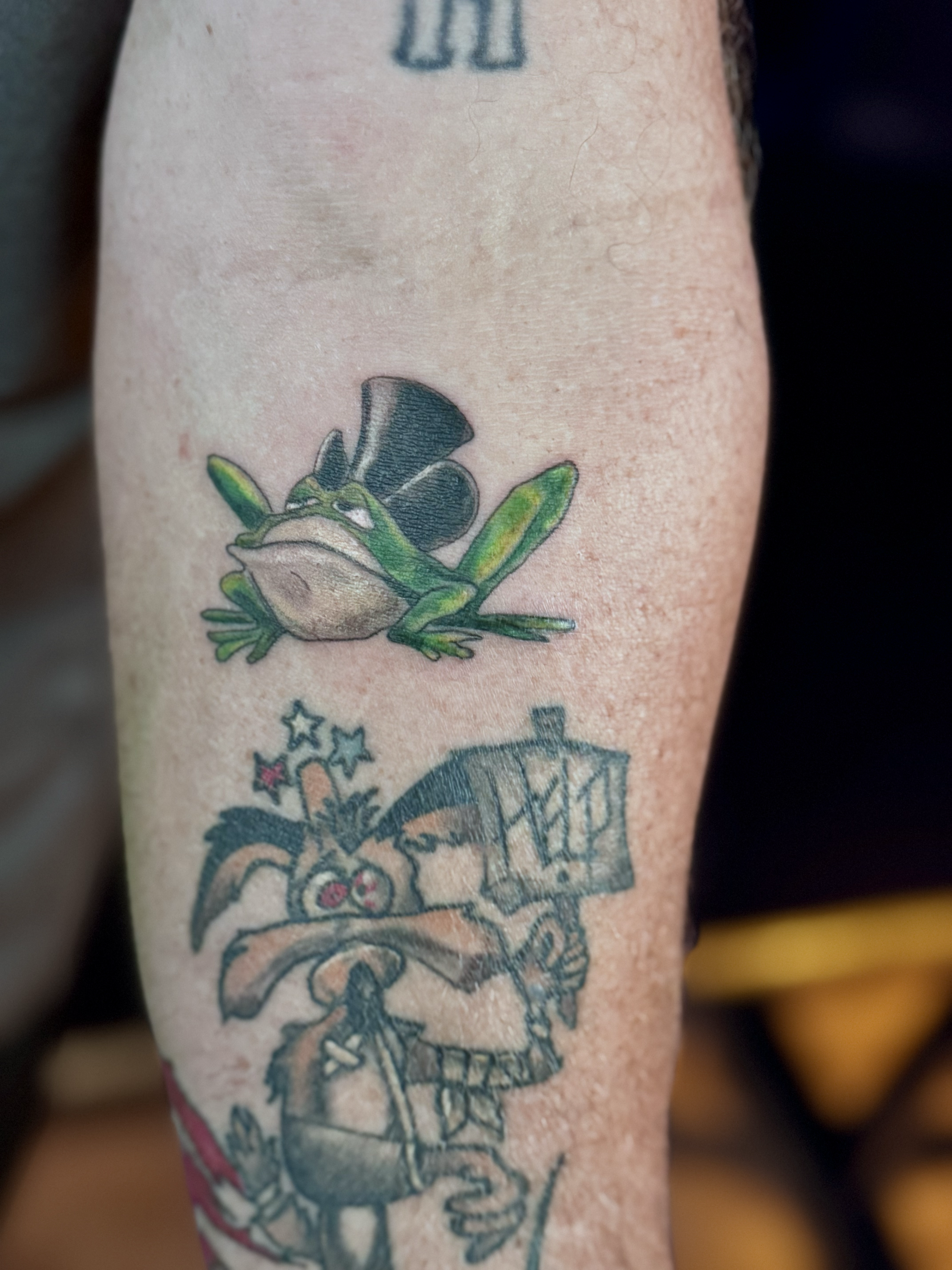 Tattoo of a frog wearing a black top hat, and a cartoonish character with bunny ears, holding a sign or book, with colorful stars around.