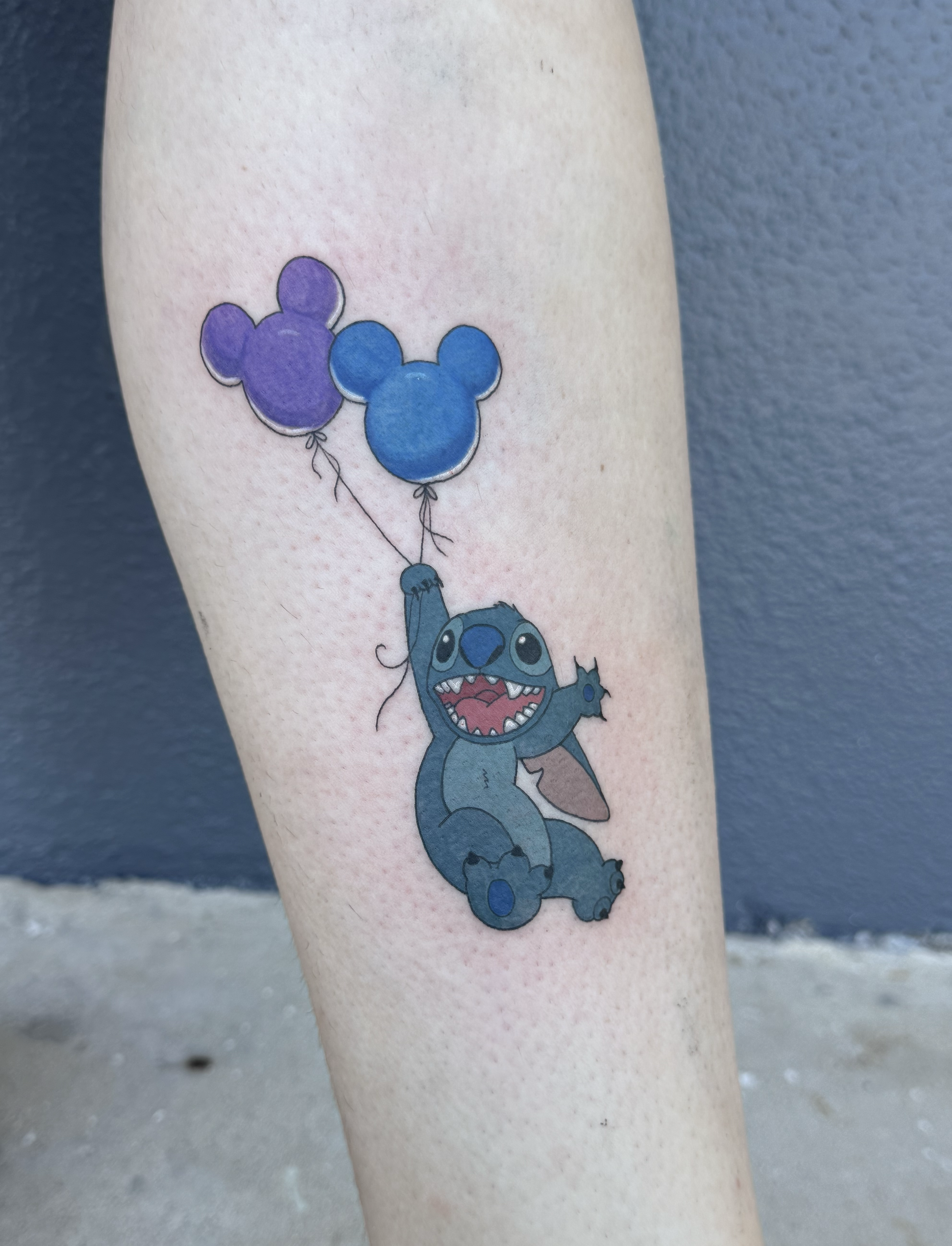 Tattoo of a cartoon alien creature holding purple and blue balloons shaped like Mickey Mouse heads.