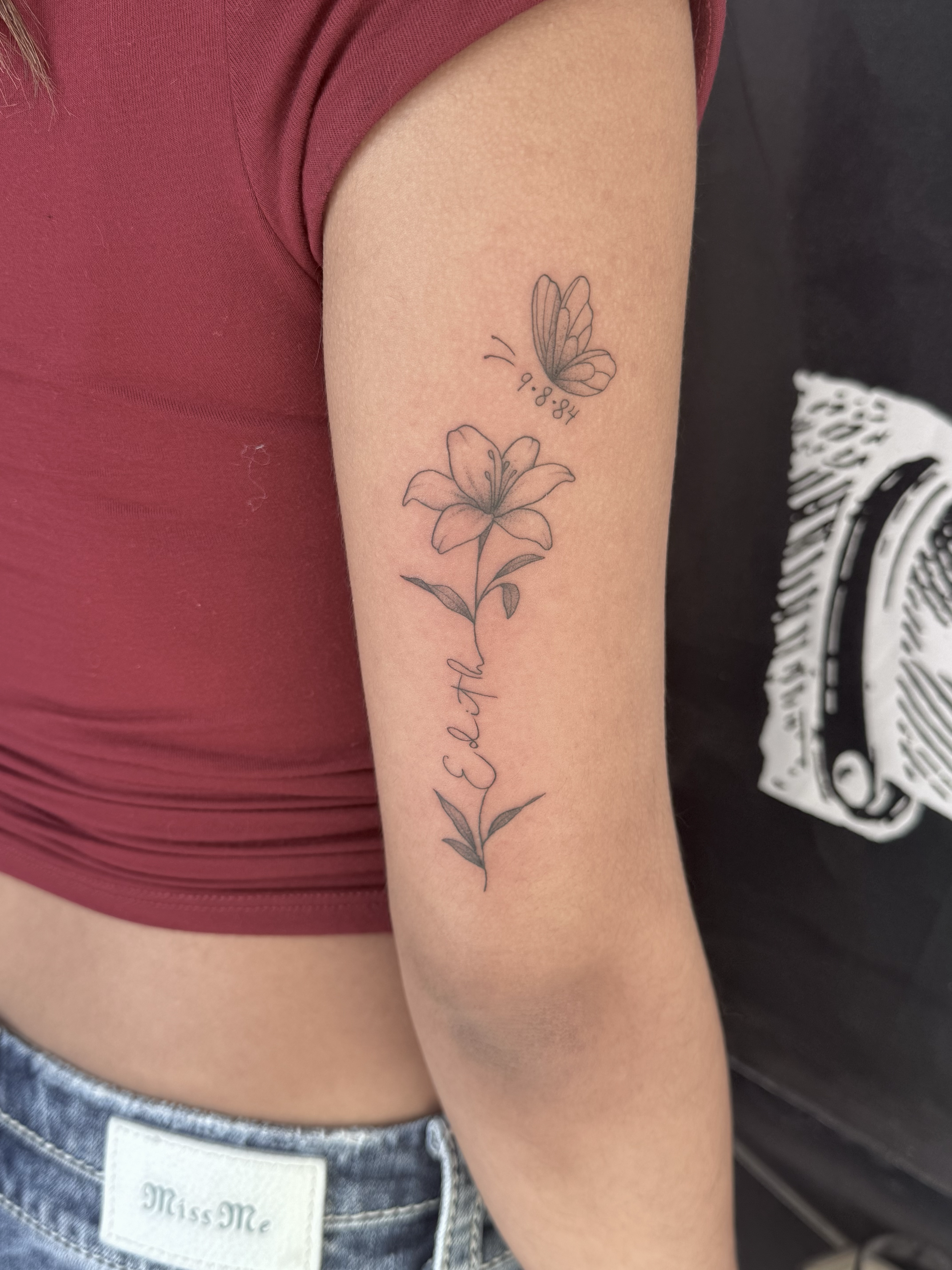 A person's upper arm with a tattoo of a flower and a butterfly. The flower is an outline of a lily with leaves, and the butterfly is outlined with details on its wings. The butterfly has the date '9.8.84' written below it, and the word 'Earth' is wri