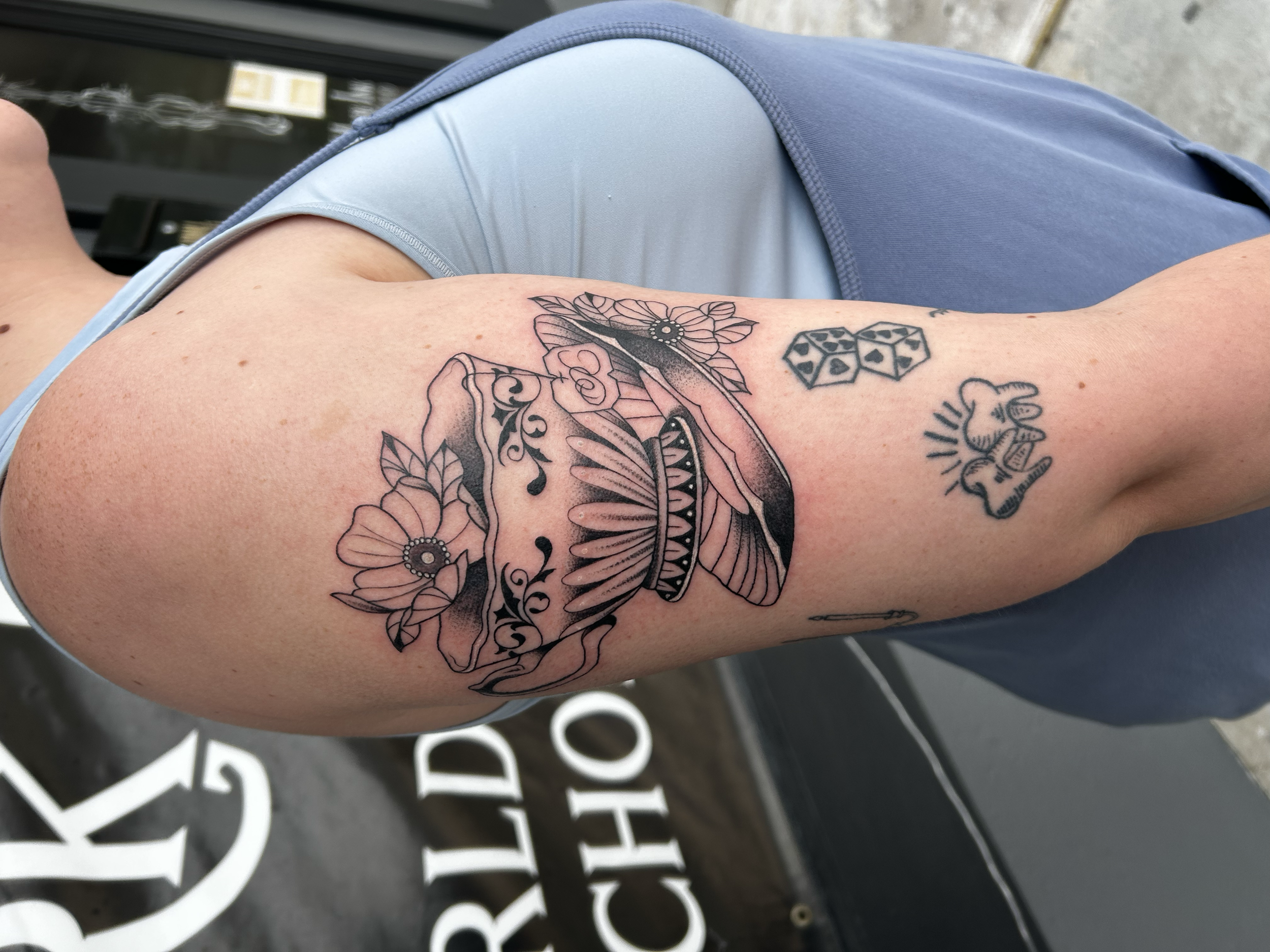 Tattoo of a traditional style pot with flowers and leaves on a person's upper arm, with additional smaller tattoos of dice, a bear paw, and a needle.
