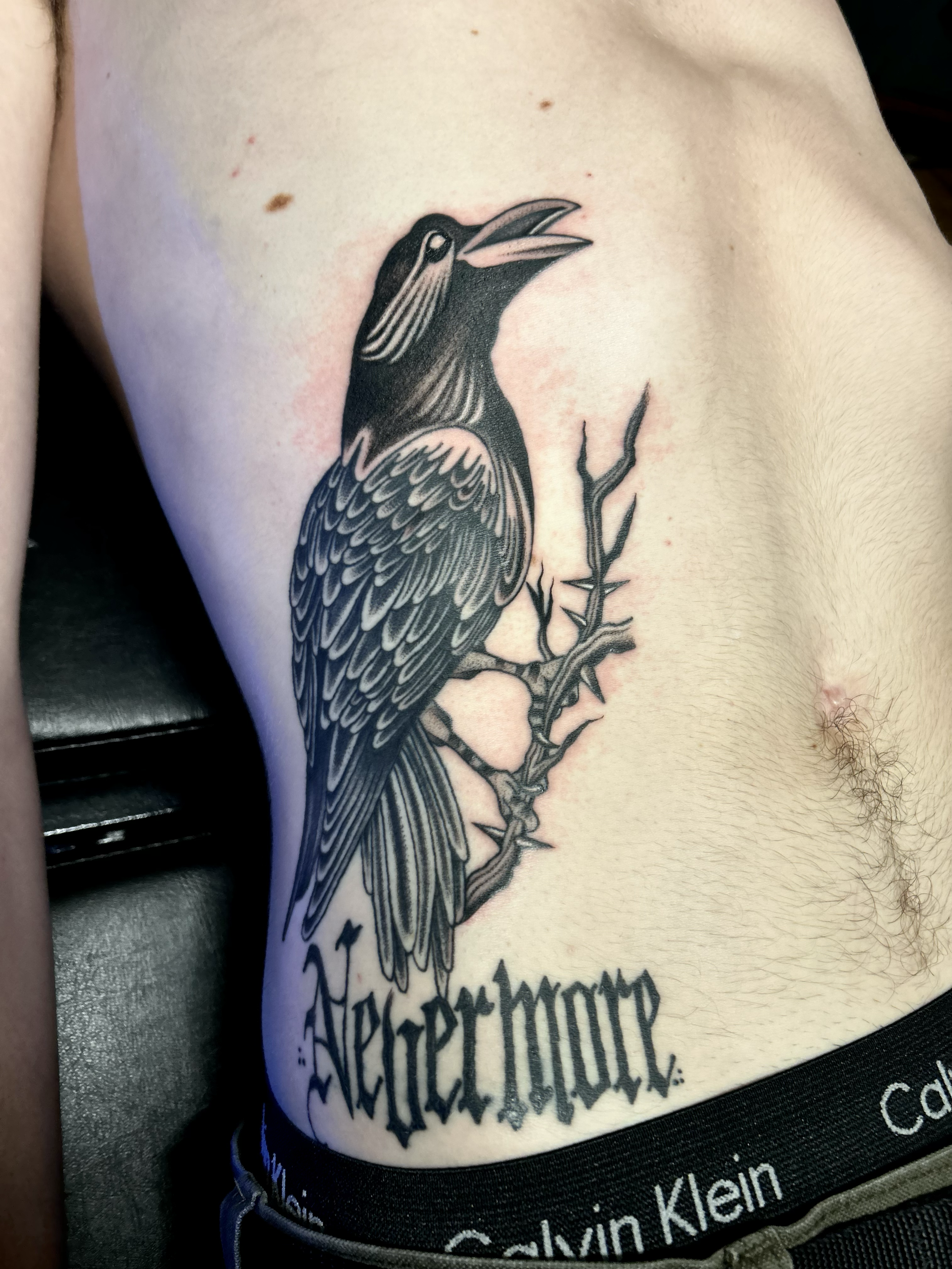 Black and gray tattoo of a crow perched on a branch, with the word 'Aperfhorr' in gothic script below, on a person's side torso.